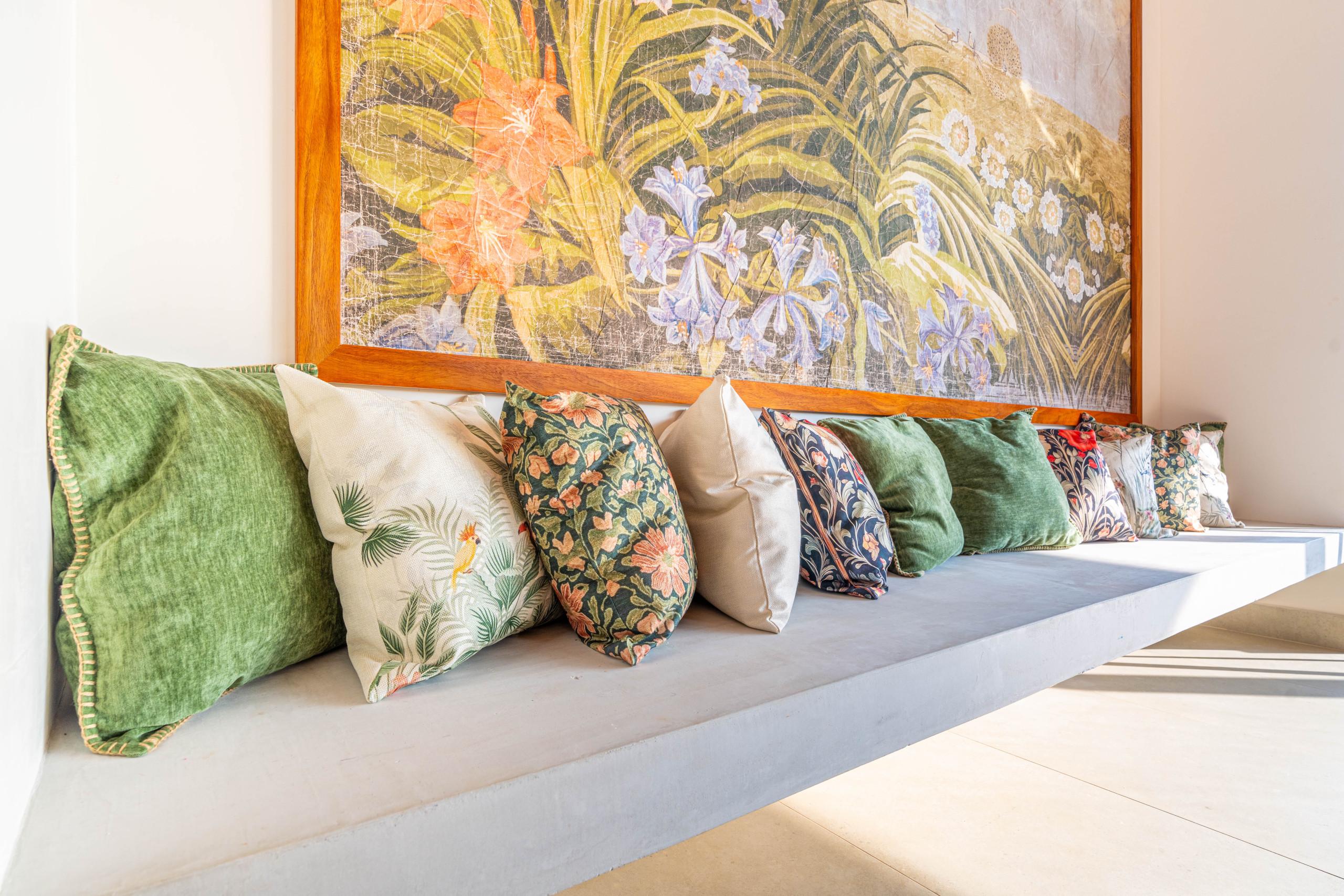 A built-in concrete bench with an array of patterned pillows sits beneath a large framed tropical artwork, bathed in natural light.