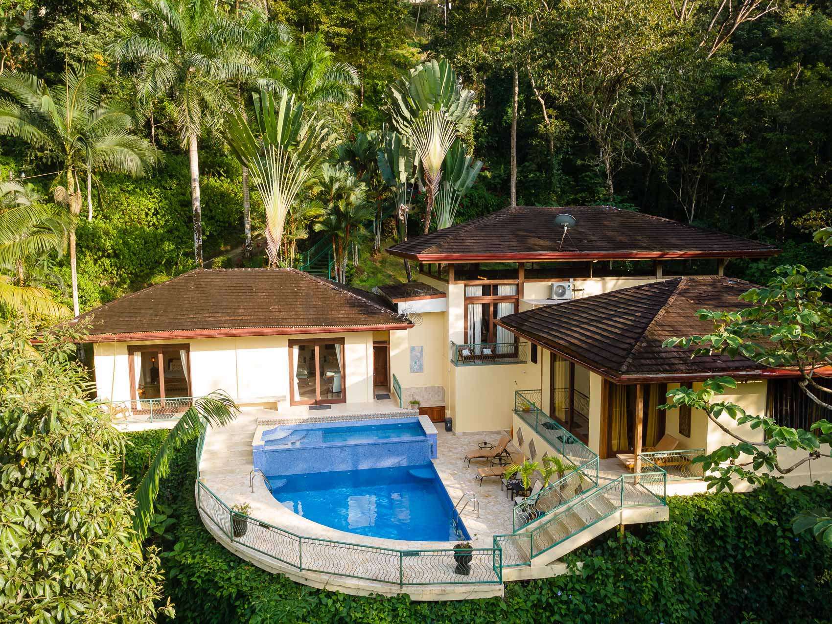 An aerial view reveals a multi-structured villa with Balinese-inspired architecture, featuring a large main pool and a separate wading pool, all nestled within a vibrant tropical jungle.