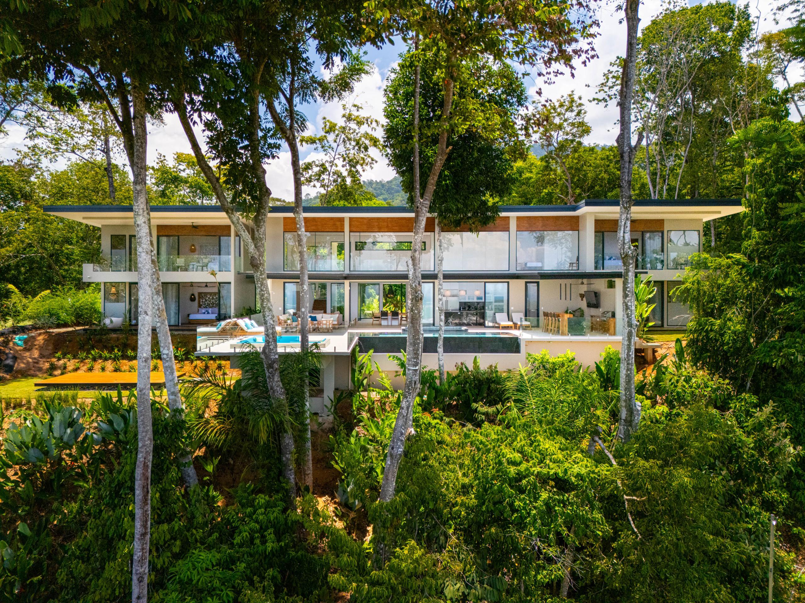 A multi-level modern villa is set into a tropical hillside, featuring an infinity pool on a wide terrace, expansive glass walls, and multiple outdoor living areas framed by lush jungle.