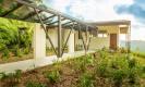 A modern, open-air walkway shaded by a translucent canopy leads through tropical landscaping to a villa entrance under a bright, sunny sky.