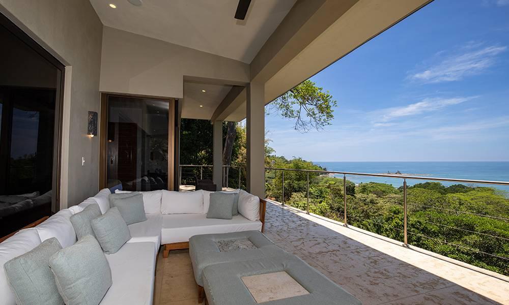 A covered outdoor living area with an L-shaped sofa opens to panoramic views of the Pacific Ocean and verdant jungle canopy.