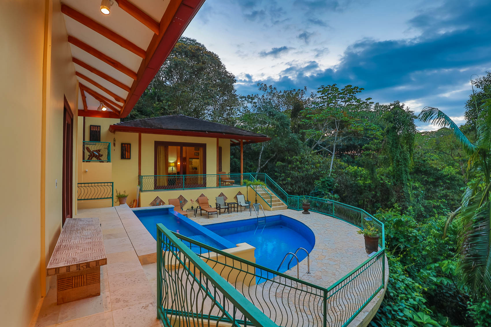 An elevated outdoor terrace features a main pool and a wading pool, bordered by tropical jungle views and Balinese-inspired architectural elements under a dramatic sky.