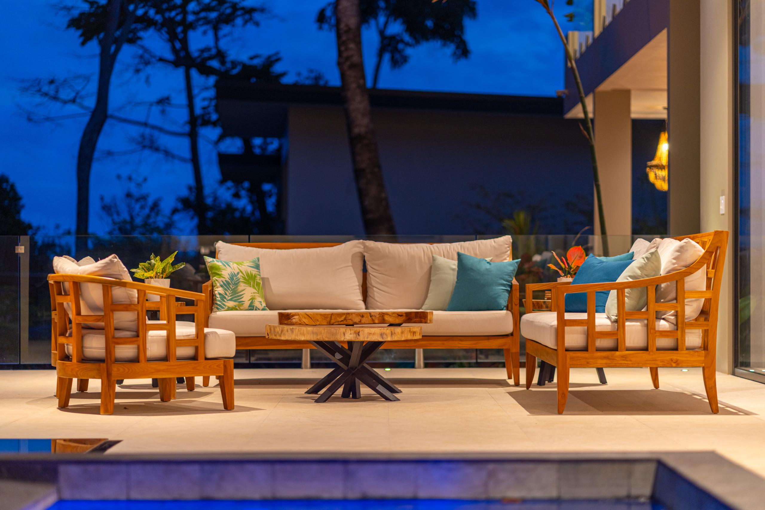A modern outdoor seating area with wooden sofas and armchairs, featuring light cushions, is set on a tiled terrace beside an illuminated pool, with a glass railing and tropical trees silhouetted against the dark evening sky.