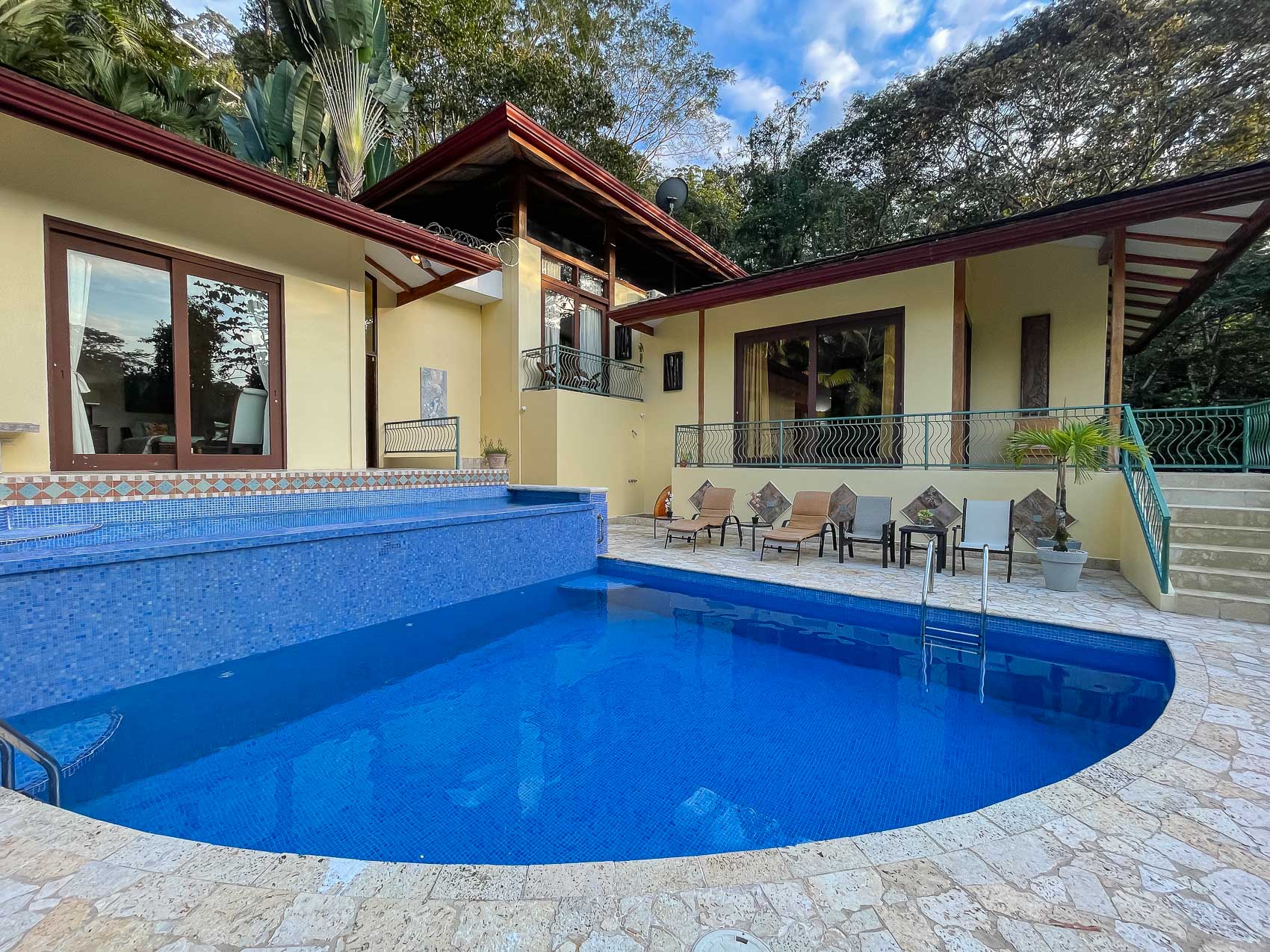 The outdoor space features a large blue tiled pool and an adjacent lounging area with tropical views, offering an inviting setting for relaxation beside the multi-level villa.