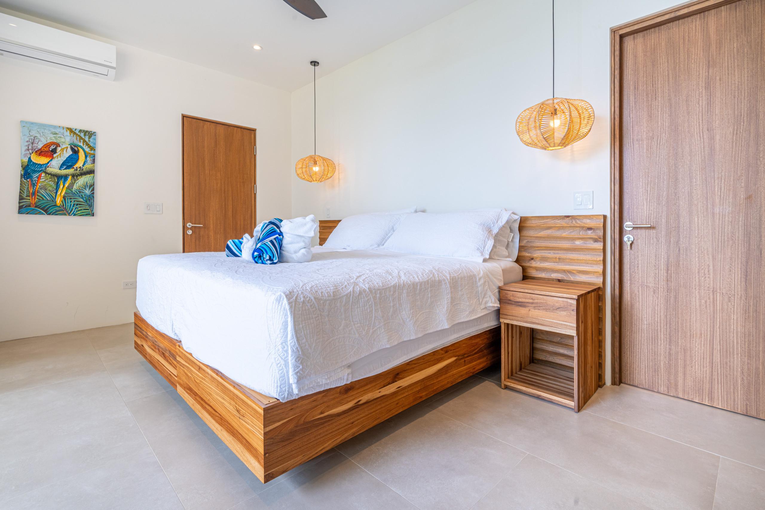 This bright bedroom features a king-size wooden bed with a slatted headboard, a matching nightstand, two woven pendant lights, light-toned walls and floors, and includes artwork and an air conditioning unit.
