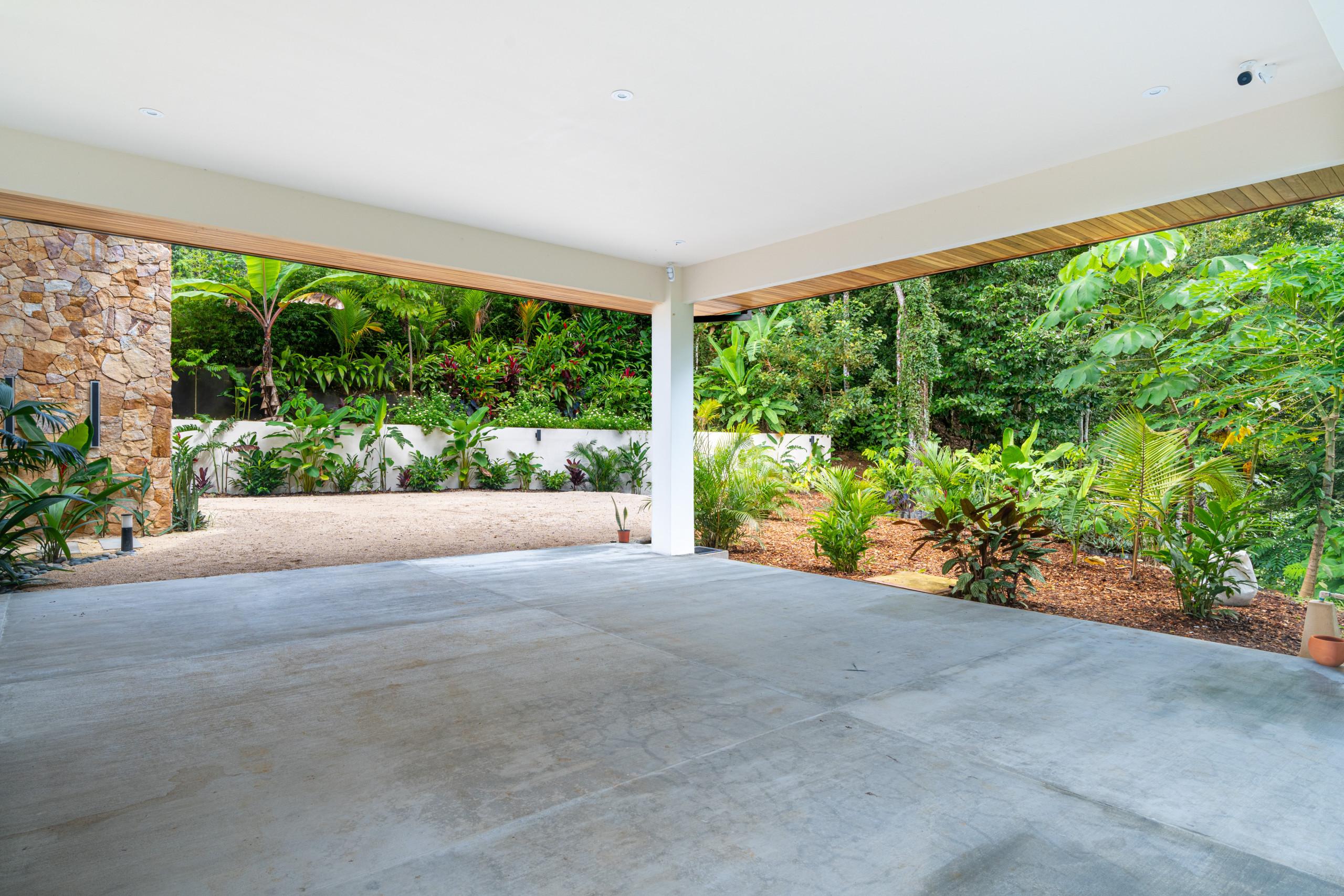 This open-air covered concrete parking area features a stone wall and timber-accented ceiling, with direct views into lush tropical gardens.