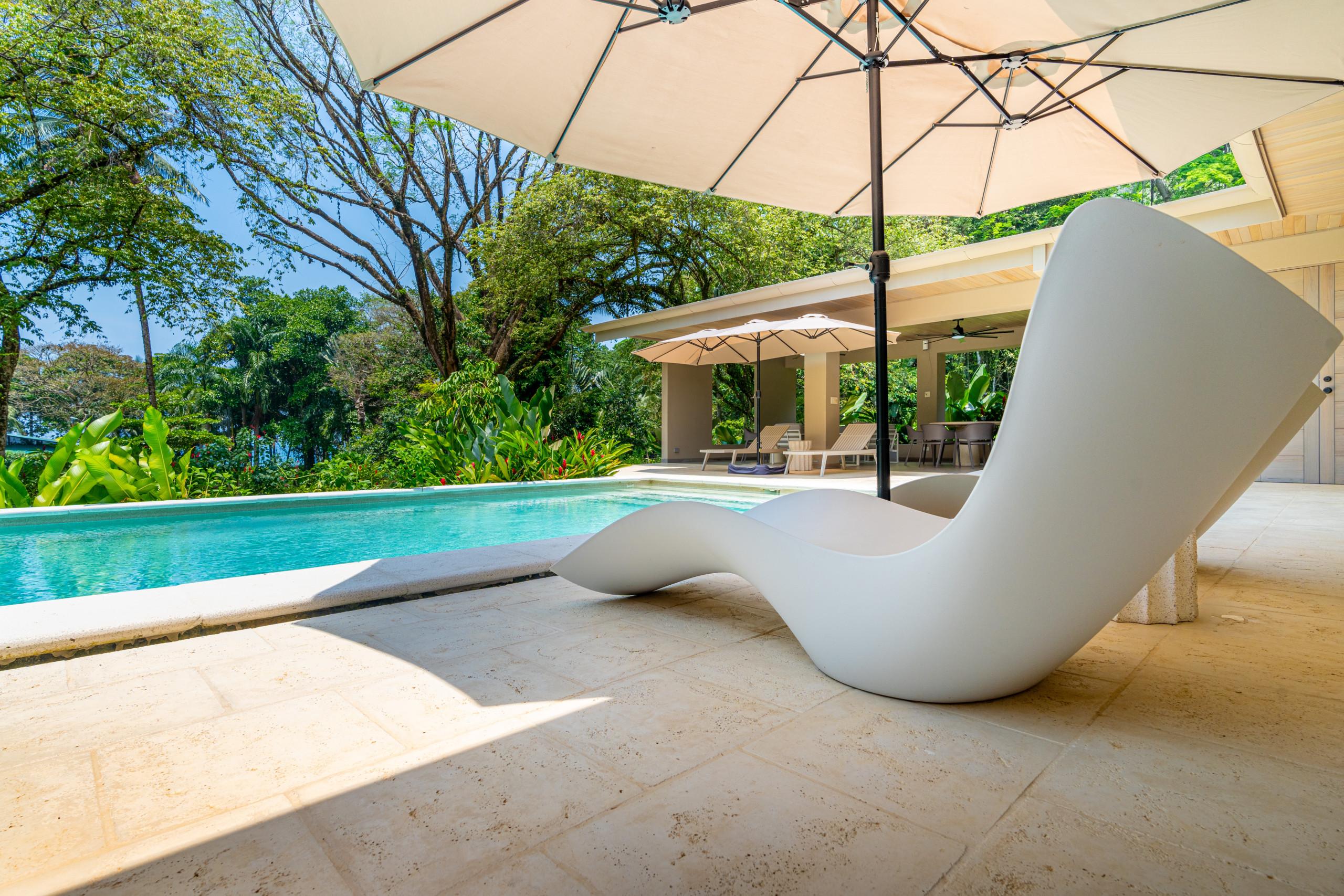 A sun-drenched outdoor terrace with light stone tiles surrounds a turquoise pool, leading to a covered lounge and dining area framed by lush tropical foliage and distant ocean views.