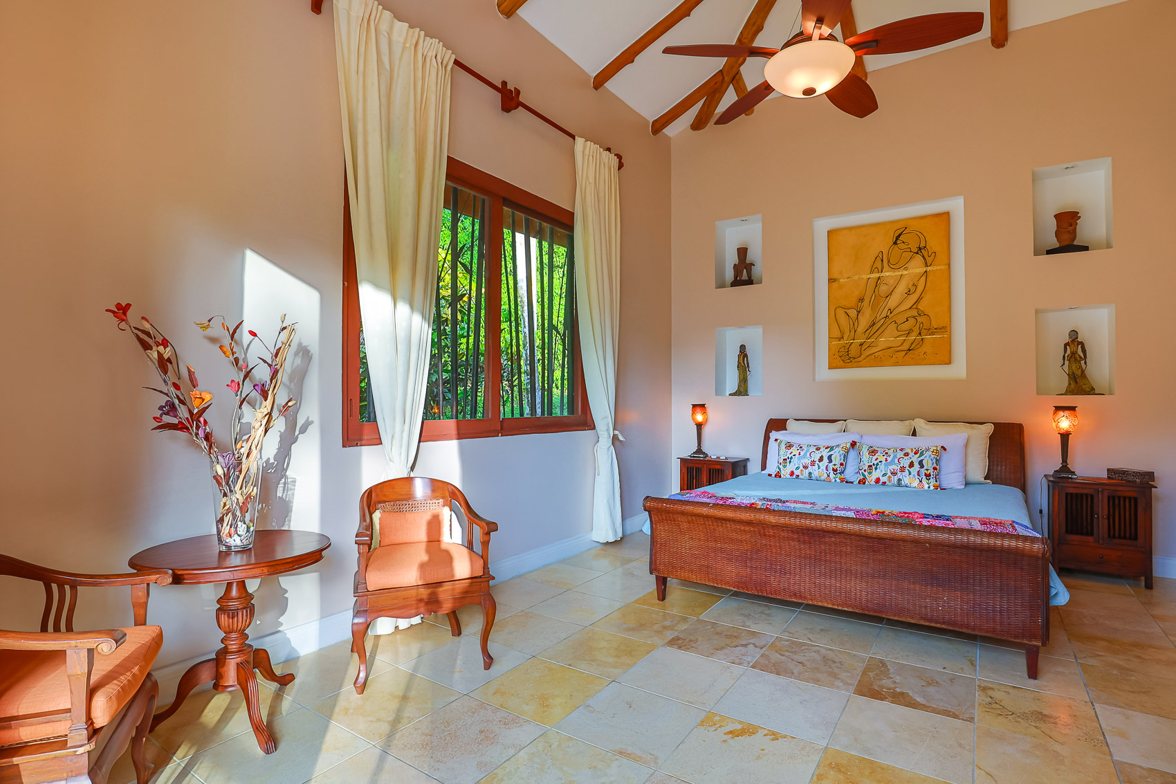 A spacious Balinese-inspired bedroom features a king-size bed, a vaulted bamboo ceiling, and abundant natural light entering through a large window with views of lush greenery.