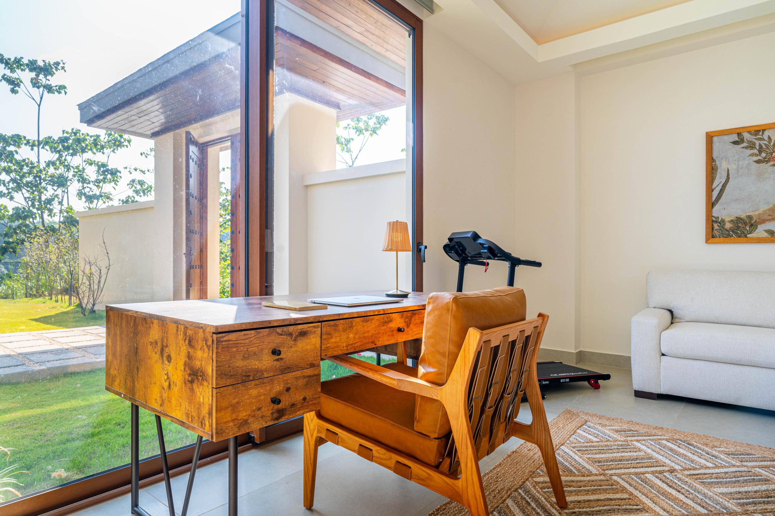 The guest suite features a wooden desk and chair, a sofa, and a treadmill, with ample natural light from large windows overlooking the tropical garden.