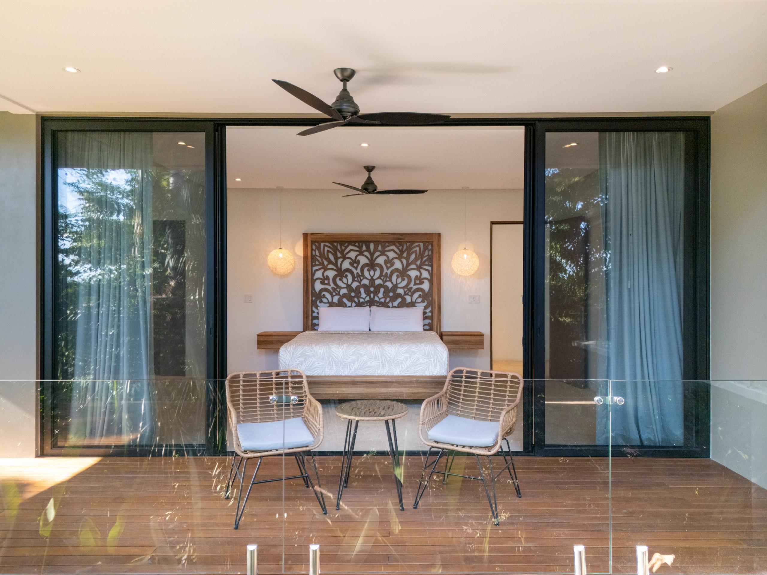 A bedroom with a king-size bed and carved wooden headboard opens through wide glass sliding doors to a private wooden terrace with outdoor seating, overlooking tropical foliage.