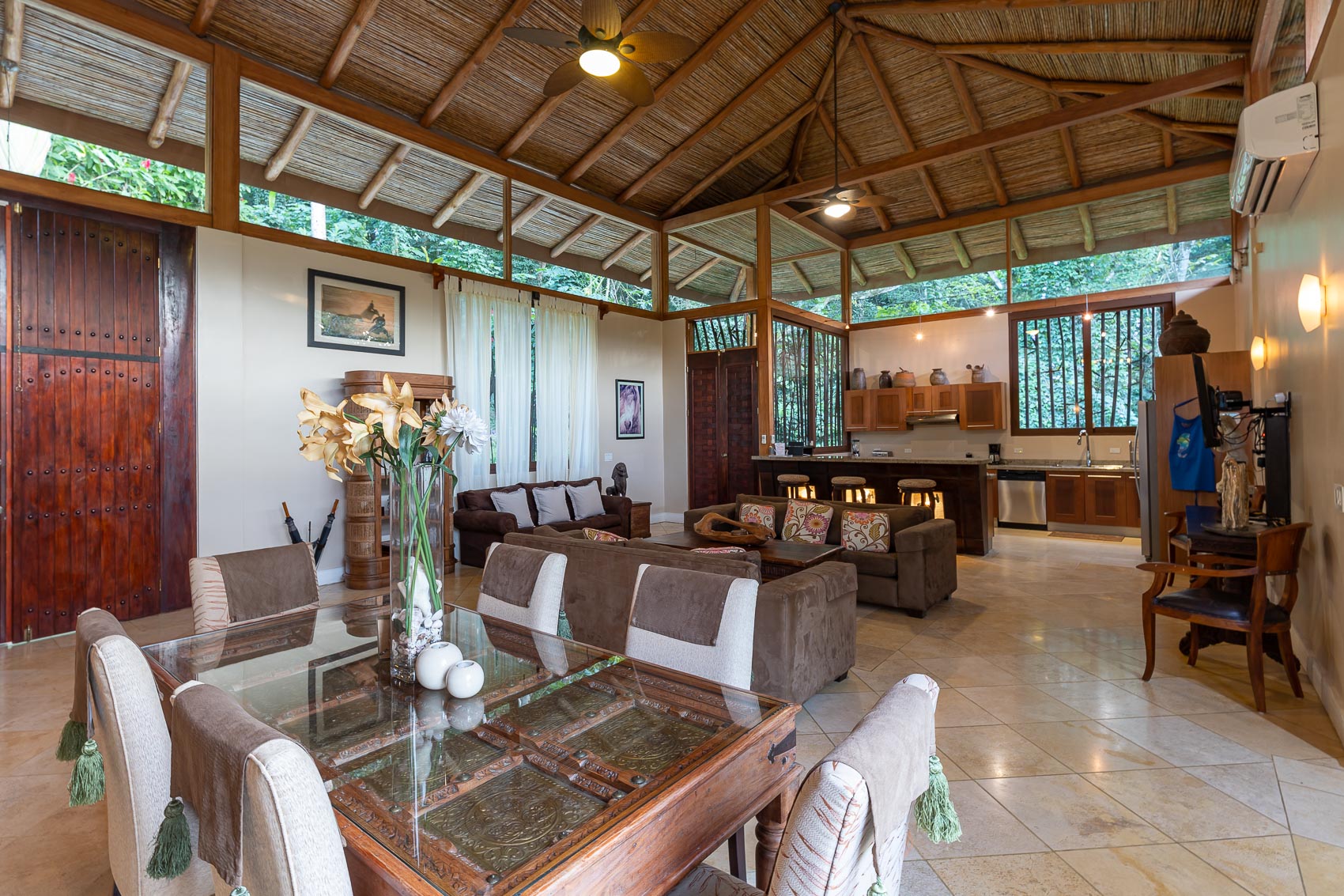 This open-plan living, dining, and kitchen space combines a high vaulted bamboo ceiling with exposed wooden beams, ample natural light, and immersive views into dense tropical foliage.