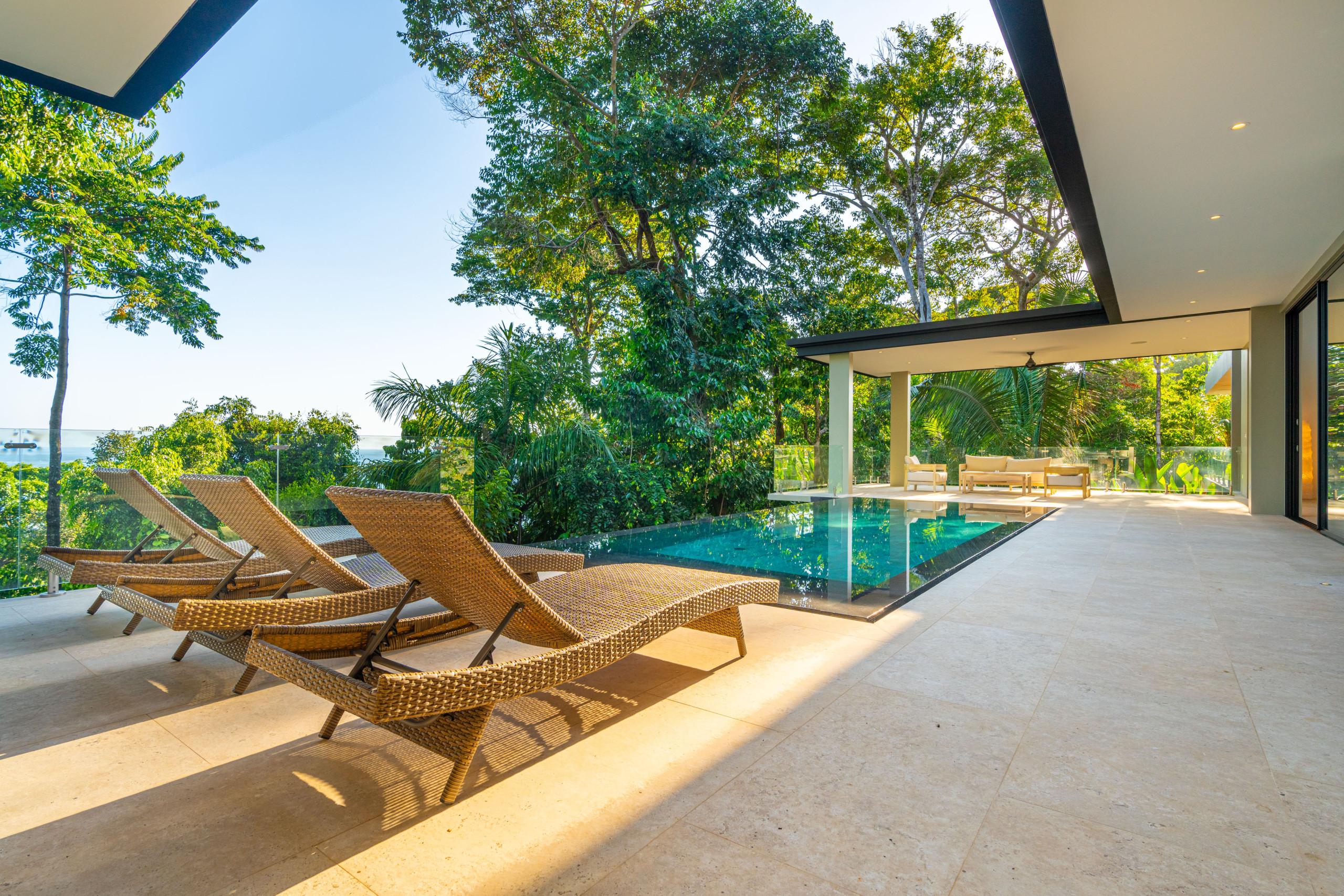 The outdoor area features an infinity pool, a spacious tiled terrace with lounge chairs and a covered seating area, all set against a backdrop of dense tropical foliage and a distant ocean view.