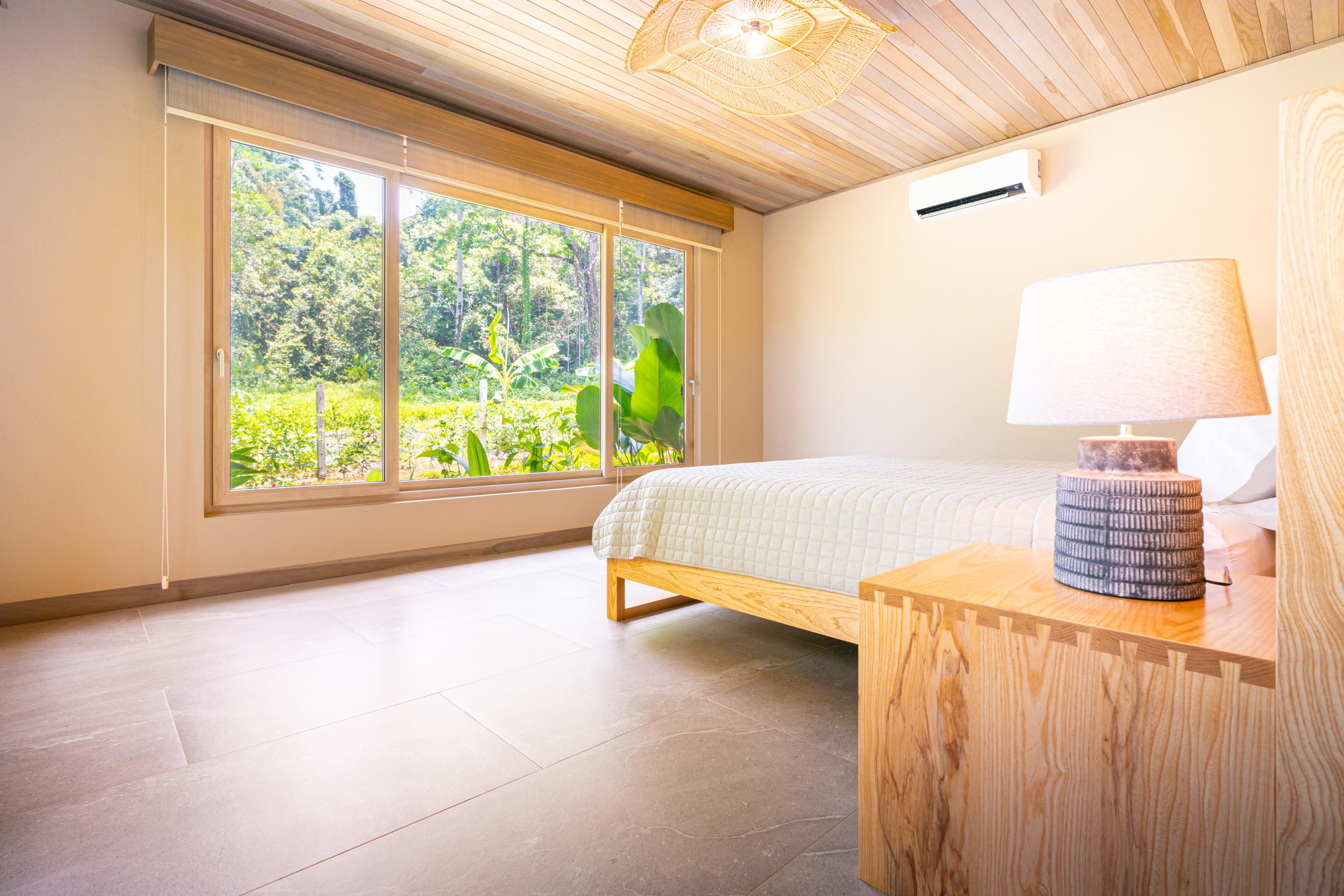 A bedroom with a light wood plank ceiling and large windows offers expansive views of the surrounding jungle, bathing the space in natural light.