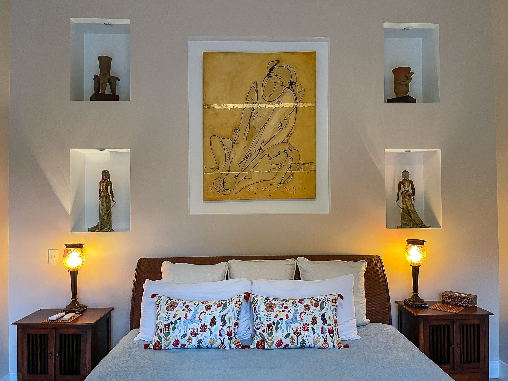 This bedroom features a king-sized bed with an ornate woven headboard, flanked by warmly lit bedside tables, and a culturally inspired wall design with a central artwork and illuminated display niches.