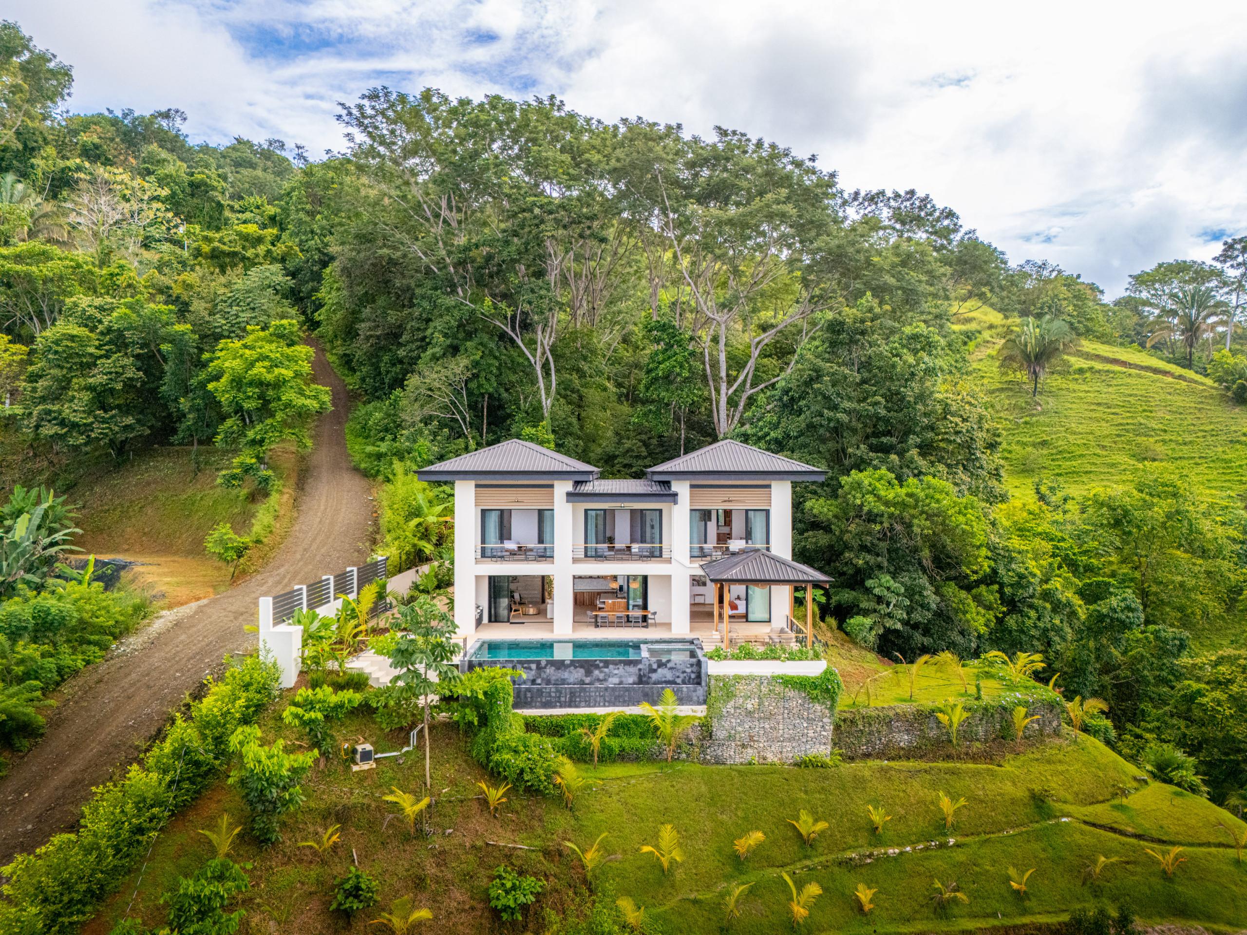 The modern two-story villa with white walls and dark roofs features an infinity pool and multiple open terraces, situated on a hillside surrounded by dense tropical jungle and landscaped gardens, accessed by a winding dirt path.