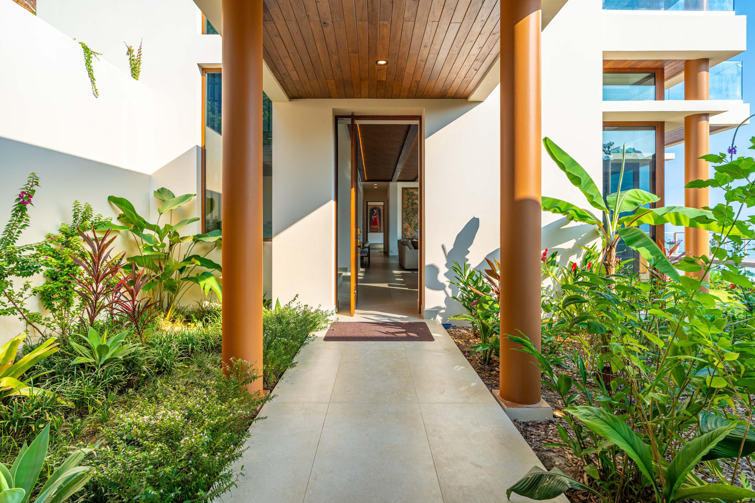 A spacious entrance features a tiled path, architectural columns, a wooden ceiling, and abundant tropical plants, opening into the home's interior with natural light and hints of the surrounding views.