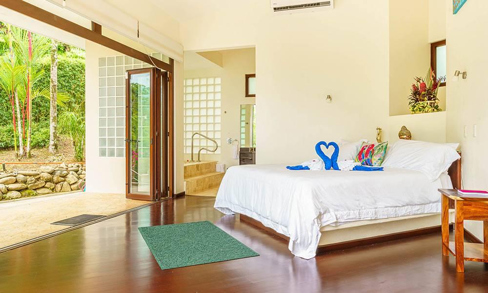 This bedroom connects directly to a lush tropical garden through wide-opening glass doors, filling the room with natural light, and offers an en-suite bathroom accessed via steps and a glass block wall.