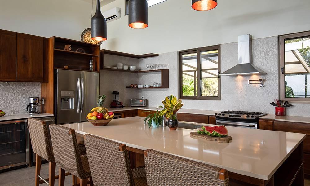 This well-lit kitchen features dark wood cabinetry, a spacious central island with bar seating, stainless steel appliances, and large windows providing views of surrounding tropical foliage.