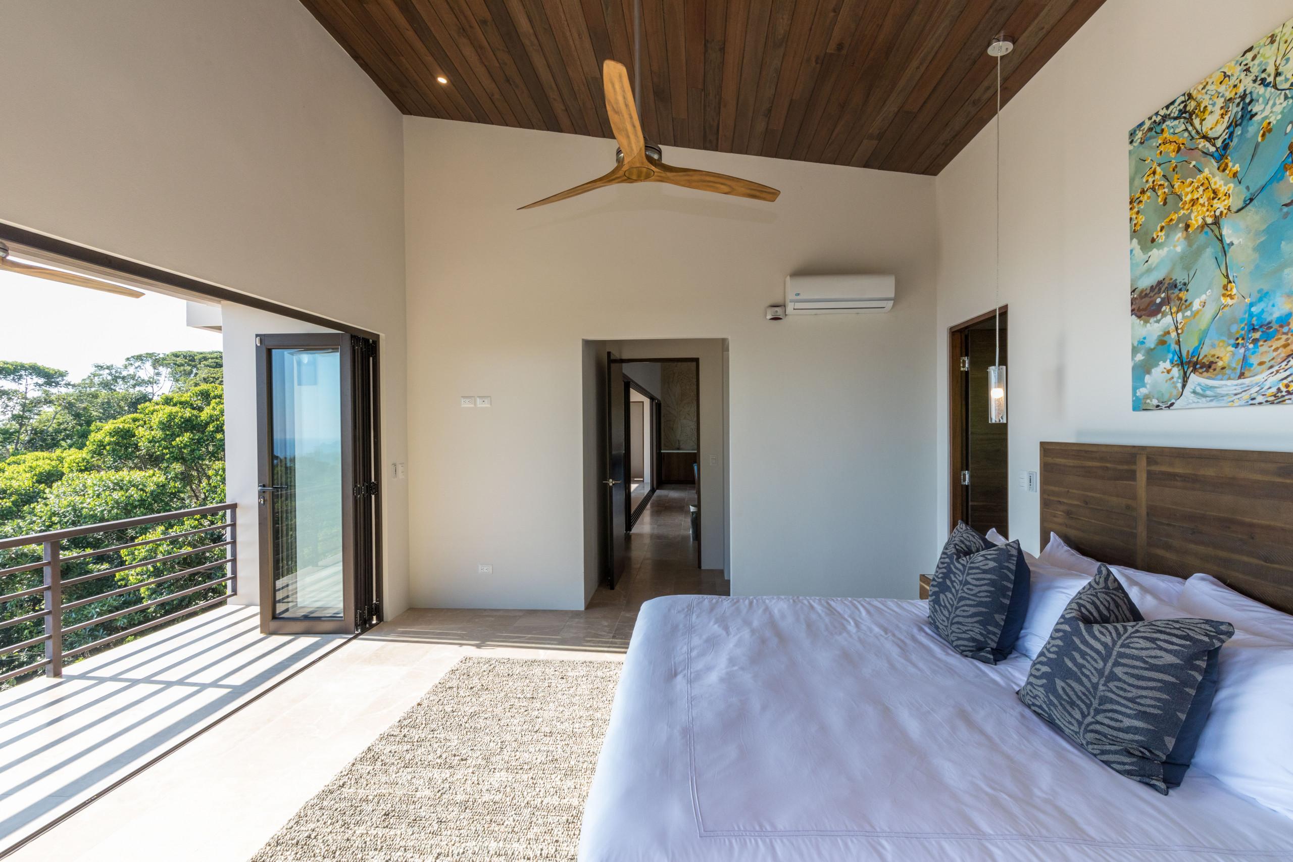 This bedroom features a king-size bed, a warm wood-paneled ceiling, and folding glass doors that provide natural light and access to a terrace overlooking the jungle.