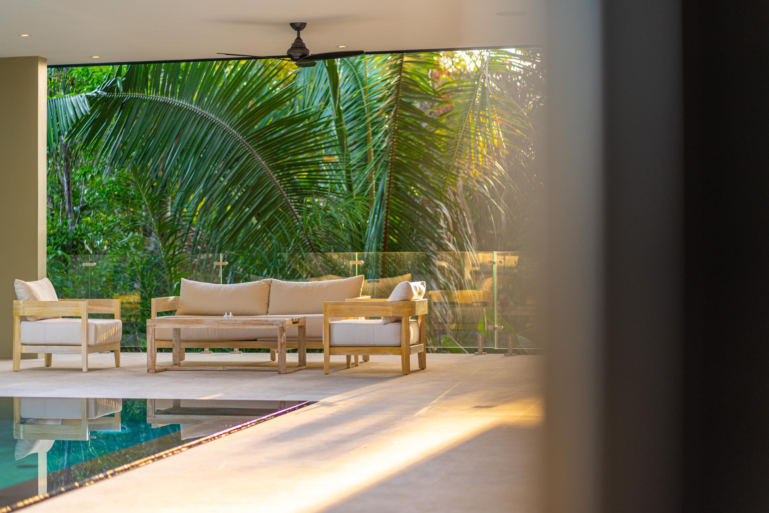 A covered terrace features a wood-framed outdoor seating area with light cushions, overlooking tropical foliage under natural sunlight, with a pool bordering the space.