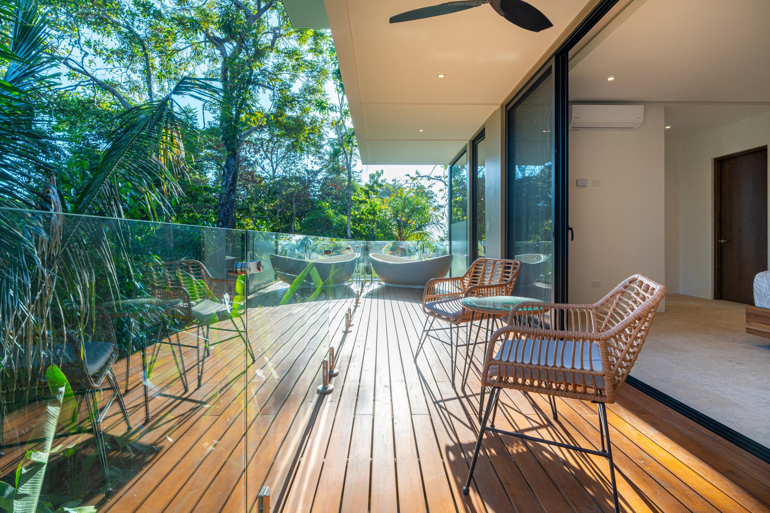 A private wooden terrace extends from a bedroom, featuring two outdoor bathtubs, a seating area with wicker chairs, and views of dense tropical jungle under natural light.