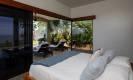 A bright bedroom with a vaulted ceiling features large glass doors opening to a private patio with lounge chairs, tropical garden, and a partial ocean view.