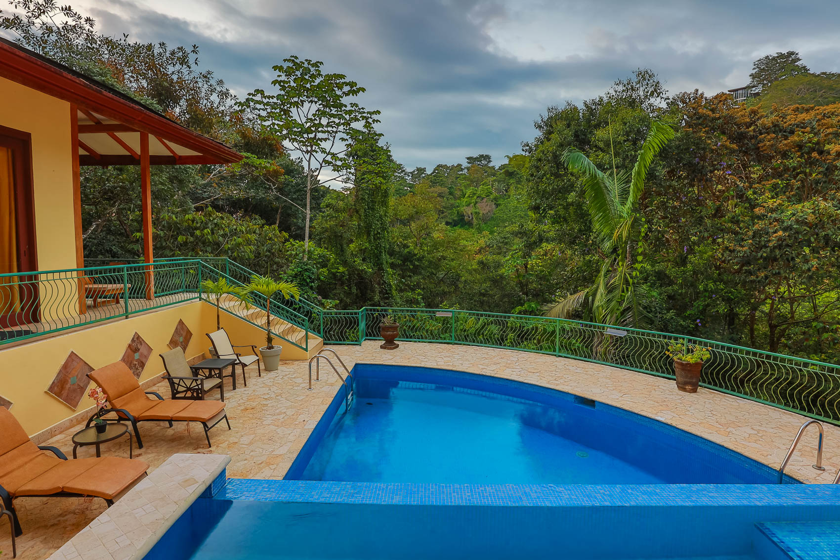 Overlooking dense tropical foliage, the villa's pool area includes a main tiled pool, a wading pool, and a stone patio with lounge seating, adjacent to a covered outdoor balcony.