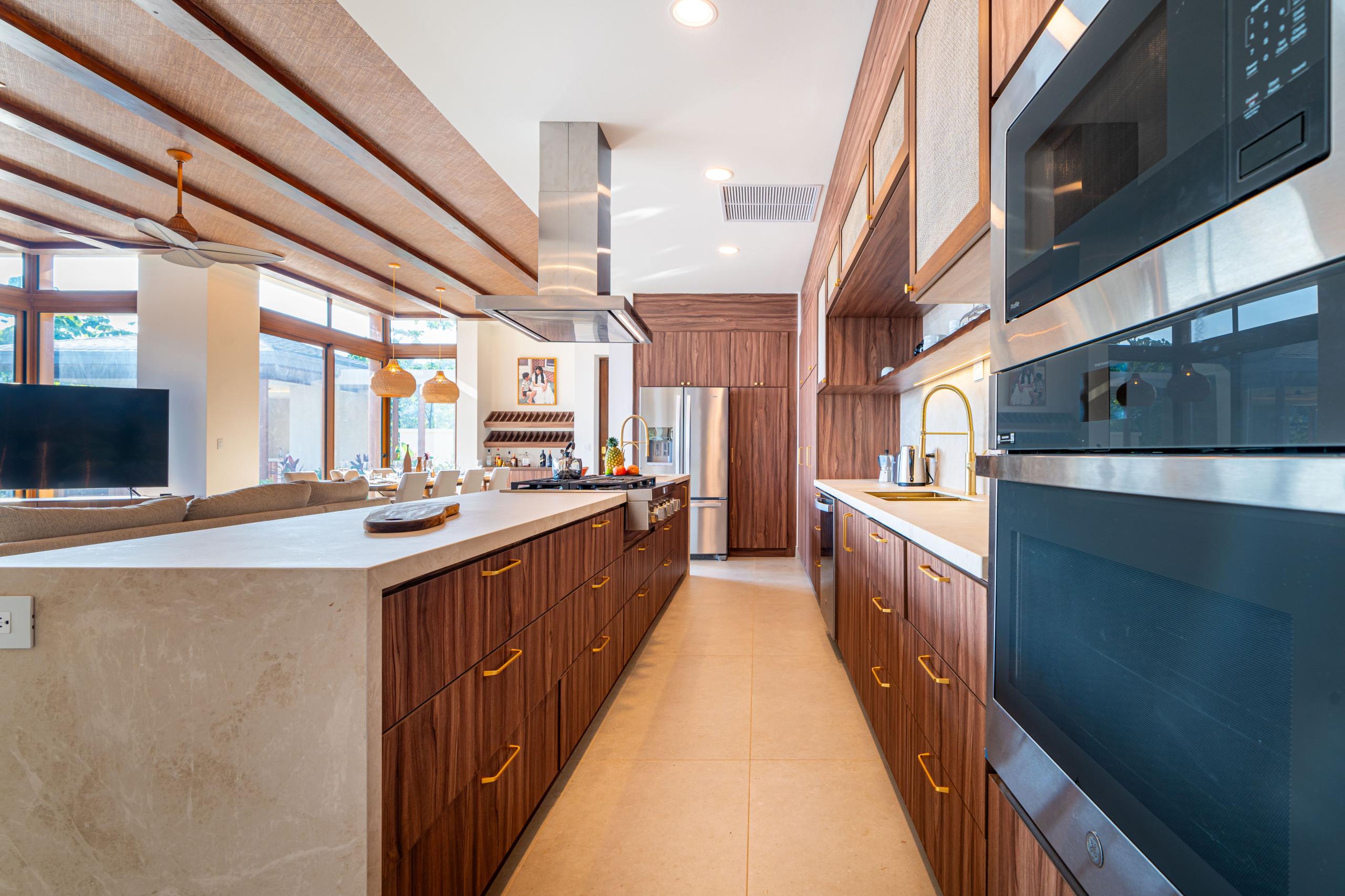This kitchen presents dark wood cabinetry, stone countertops, and stainless steel appliances, with a textured woven ceiling and large windows that provide natural light and views of the tropical environment.