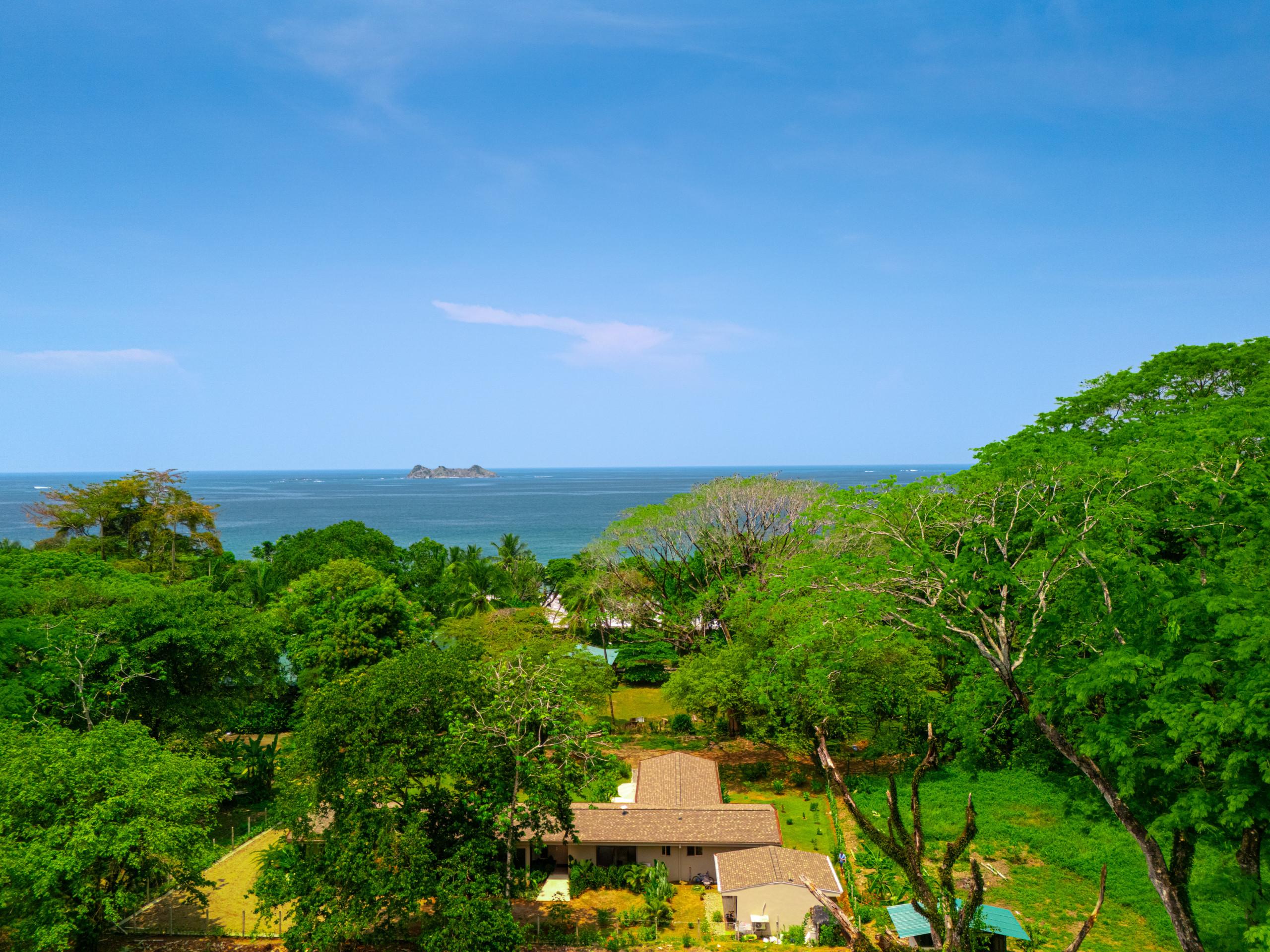 From a high vantage point, the villa's light-colored roofs peek through vibrant green jungle, offering expansive views of the calm blue ocean and a distant island.