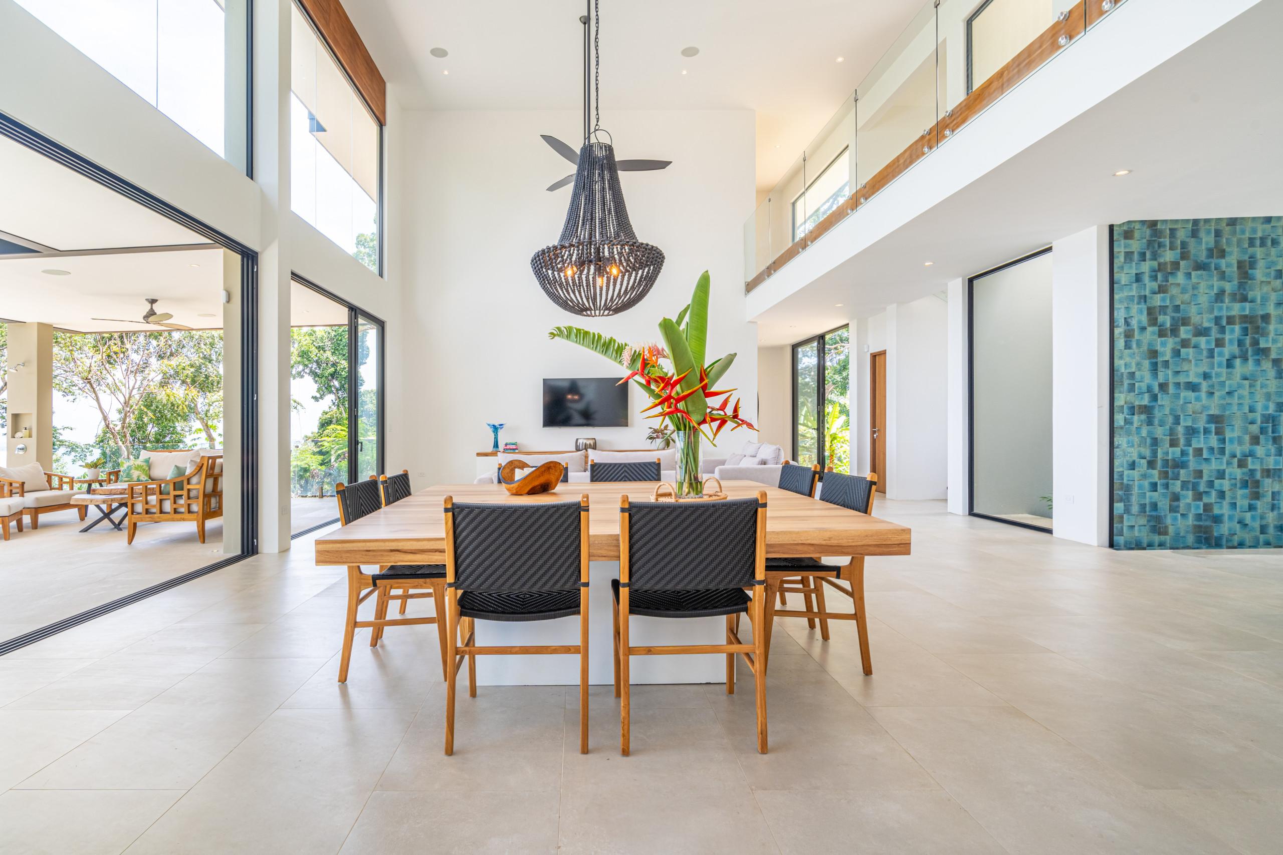 A spacious, open-concept dining room with high ceilings and extensive natural light connects directly to an outdoor terrace, offering views of tropical greenery, and is overlooked by a second-story interior balcony.