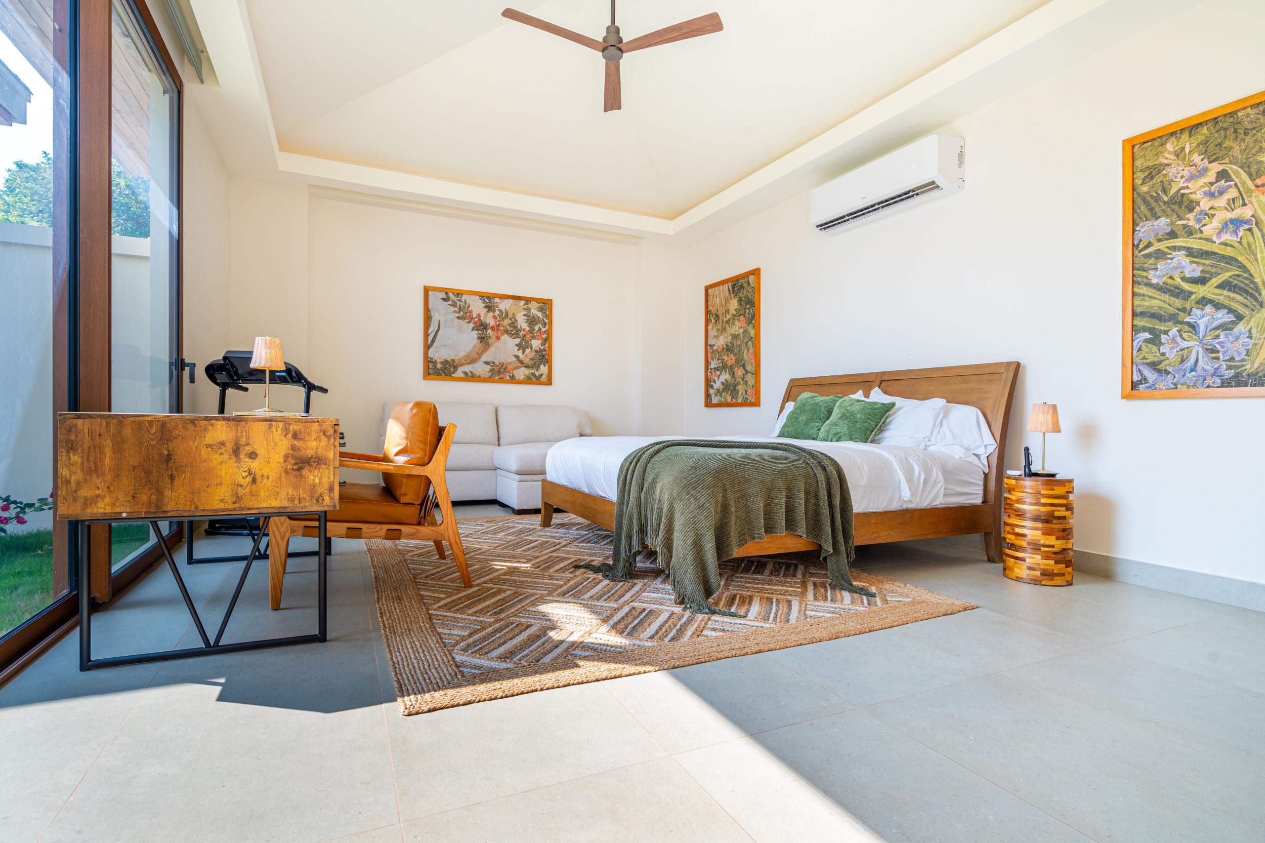 A bright guest bedroom features a king bed, a sofa, and a working desk, with large glass doors providing abundant natural light and views of the surrounding tropical landscape.