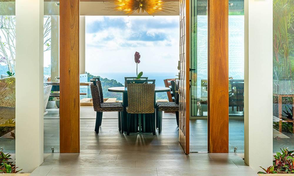 An open dining area with a circular table and woven chairs provides expansive views of the Pacific Ocean and surrounding tropical landscape.