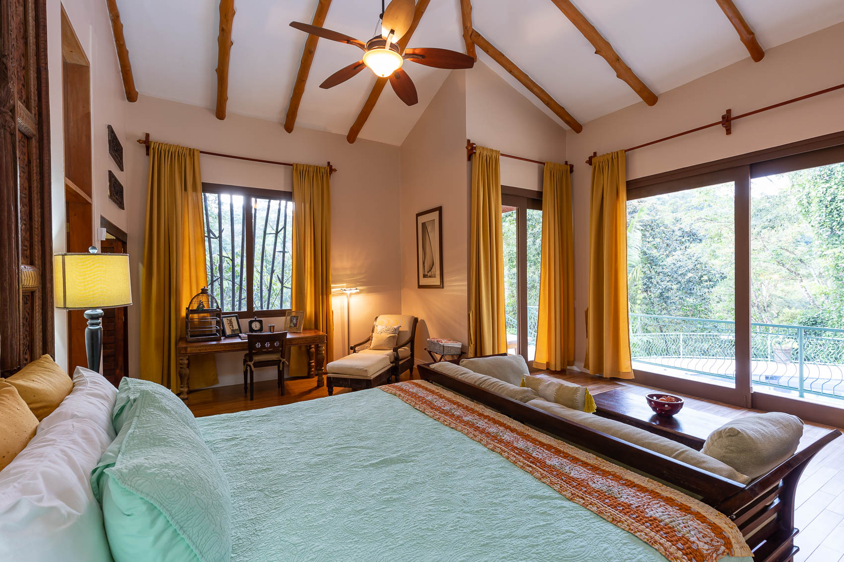 A spacious bedroom with a king bed, vaulted ceiling, and exposed wood beams features ample natural light from large sliding glass doors that open to a balcony overlooking a tropical canopy.