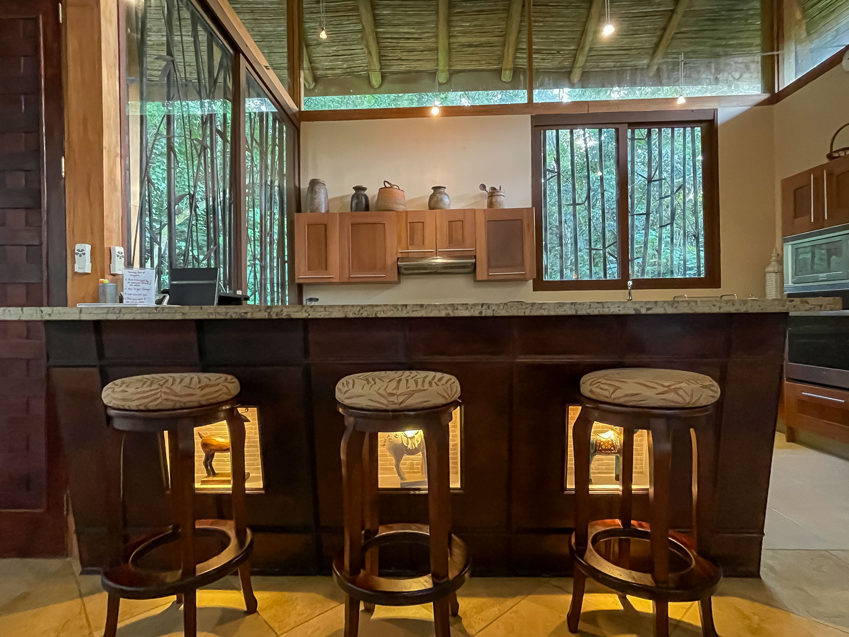 A Balinese-inspired kitchen bar with three stools and wooden cabinetry features raised bamboo ceilings and large windows that provide natural light and views of dense tropical foliage.