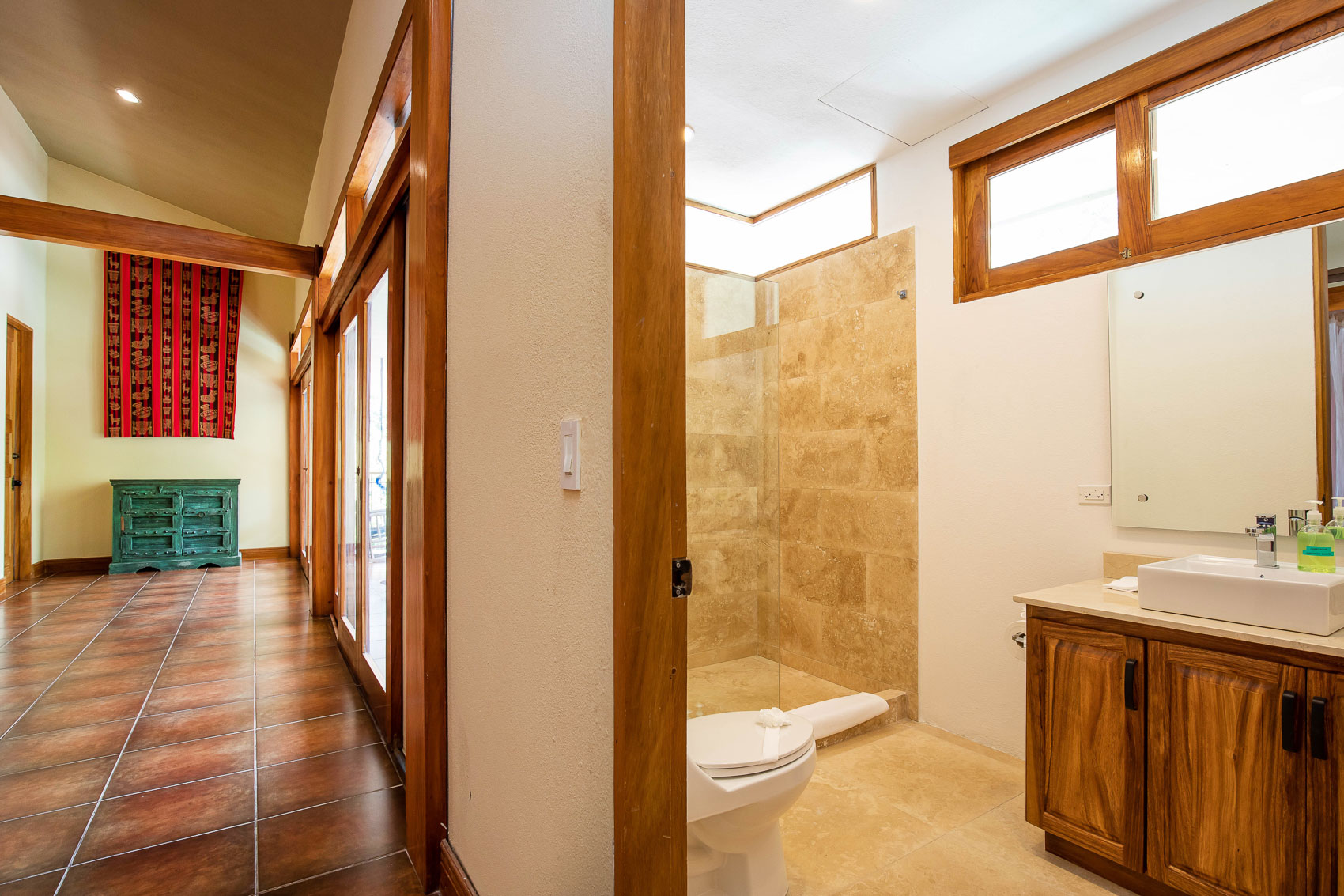 The bathroom features a spacious tiled walk-in shower and a wooden vanity, illuminated by natural light from transom windows, and connects to a hallway with large glass doors.