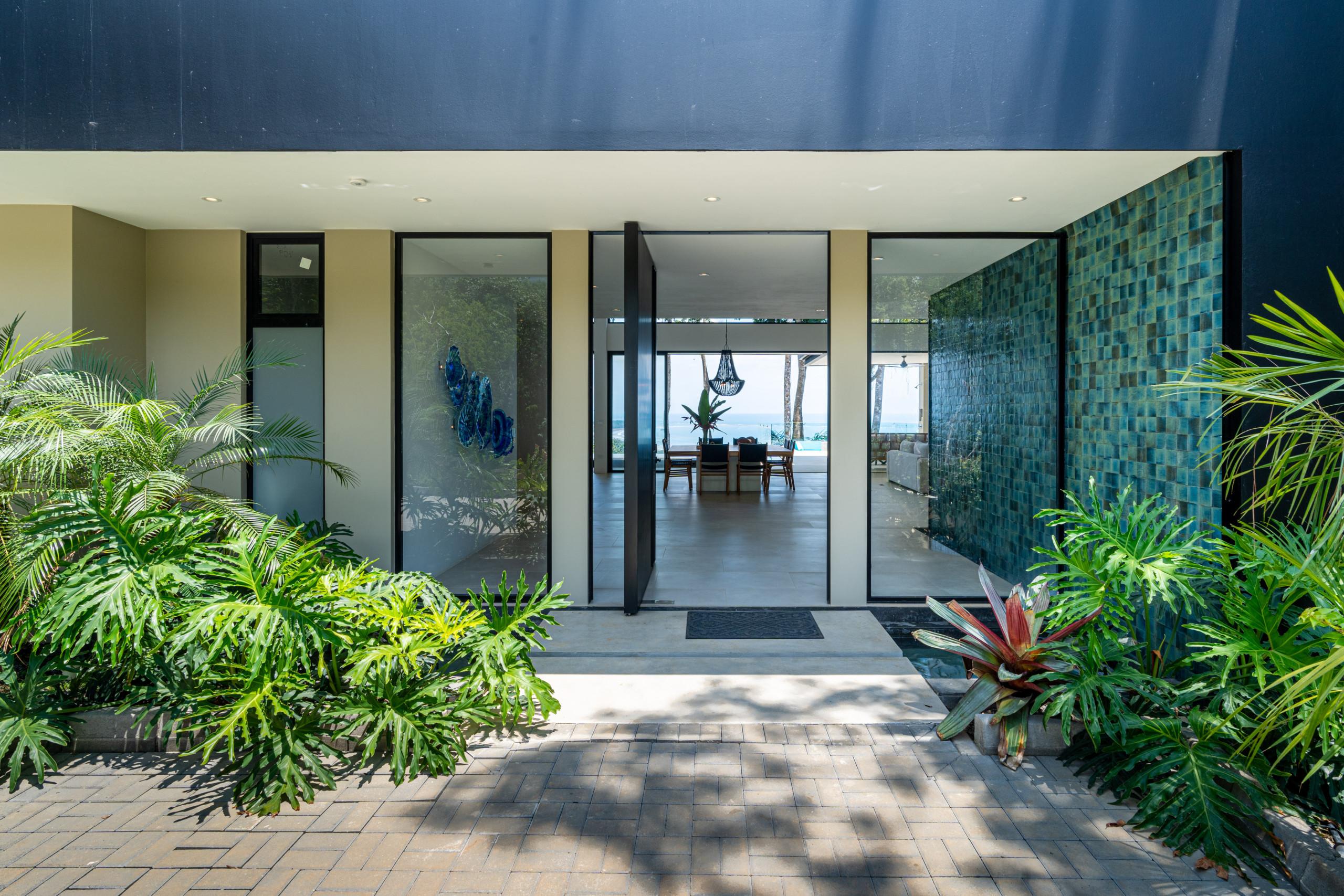 The entrance, featuring an open pivot door and expansive glass, offers immediate views of the ocean through the bright interior living and dining spaces, surrounded by tropical landscaping.