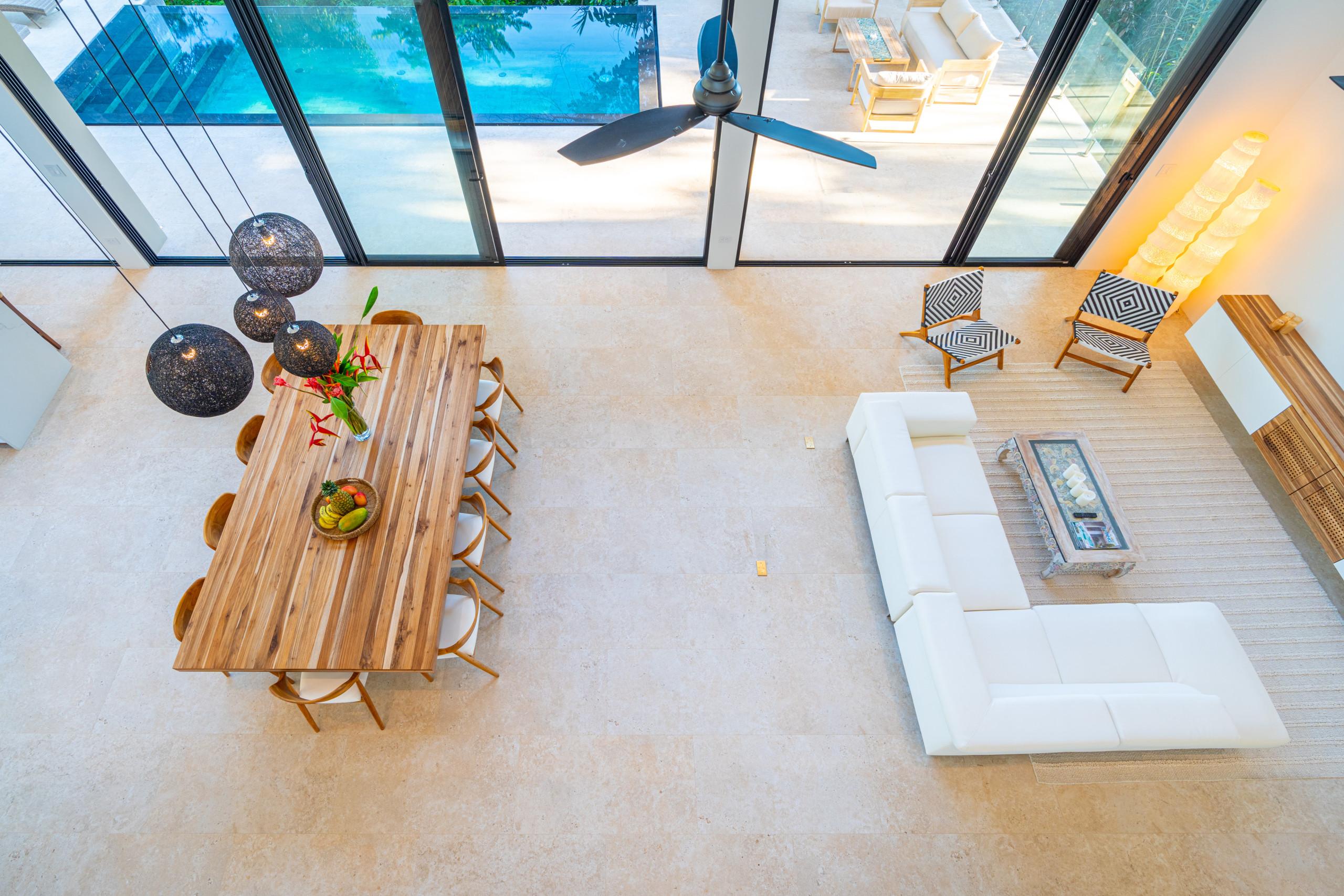 This spacious, naturally lit open-plan room features a large dining table for ten, a comfortable sectional sofa, and expansive glass walls that offer views of the outdoor pool and terrace.