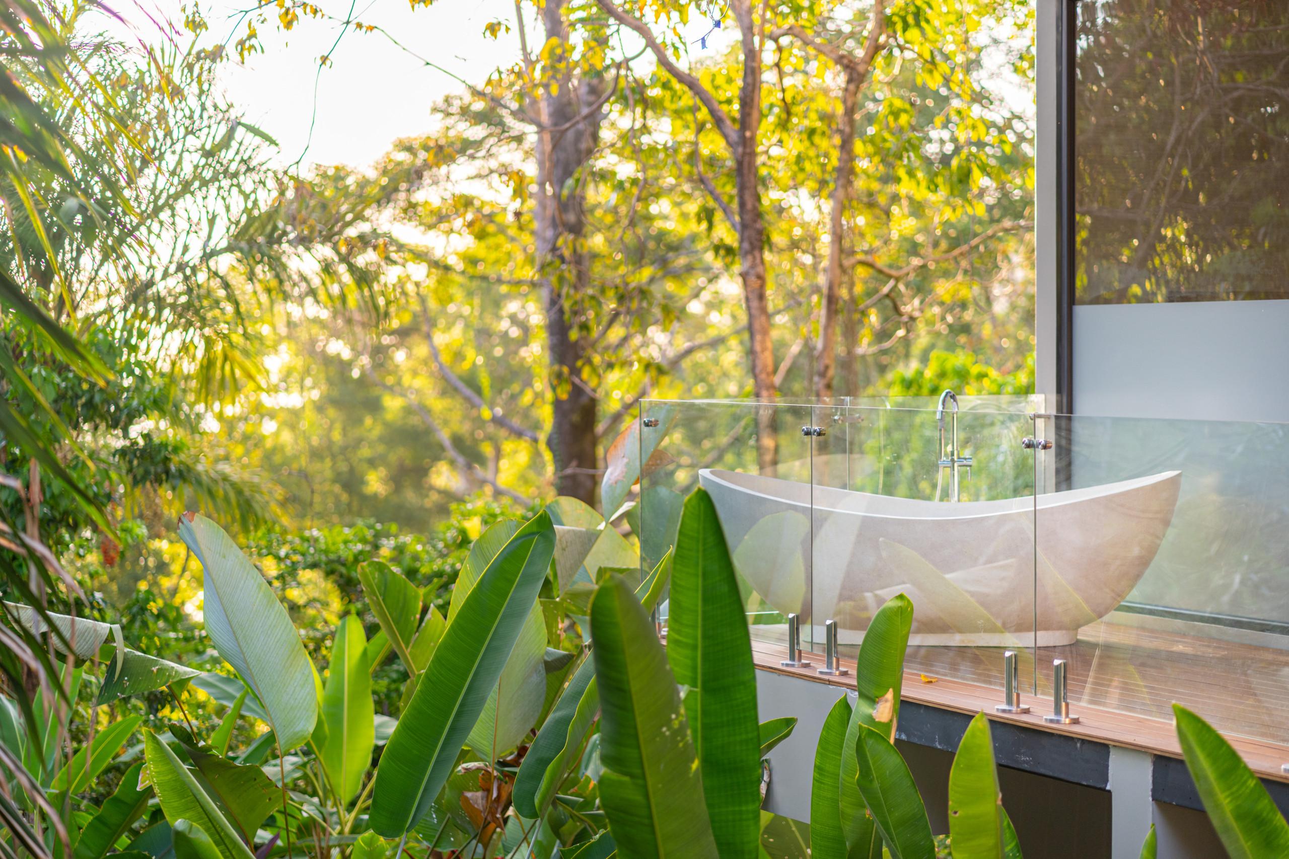 A freestanding bathtub rests on a wooden terrace with a glass railing, offering a view of sunlit tropical foliage.