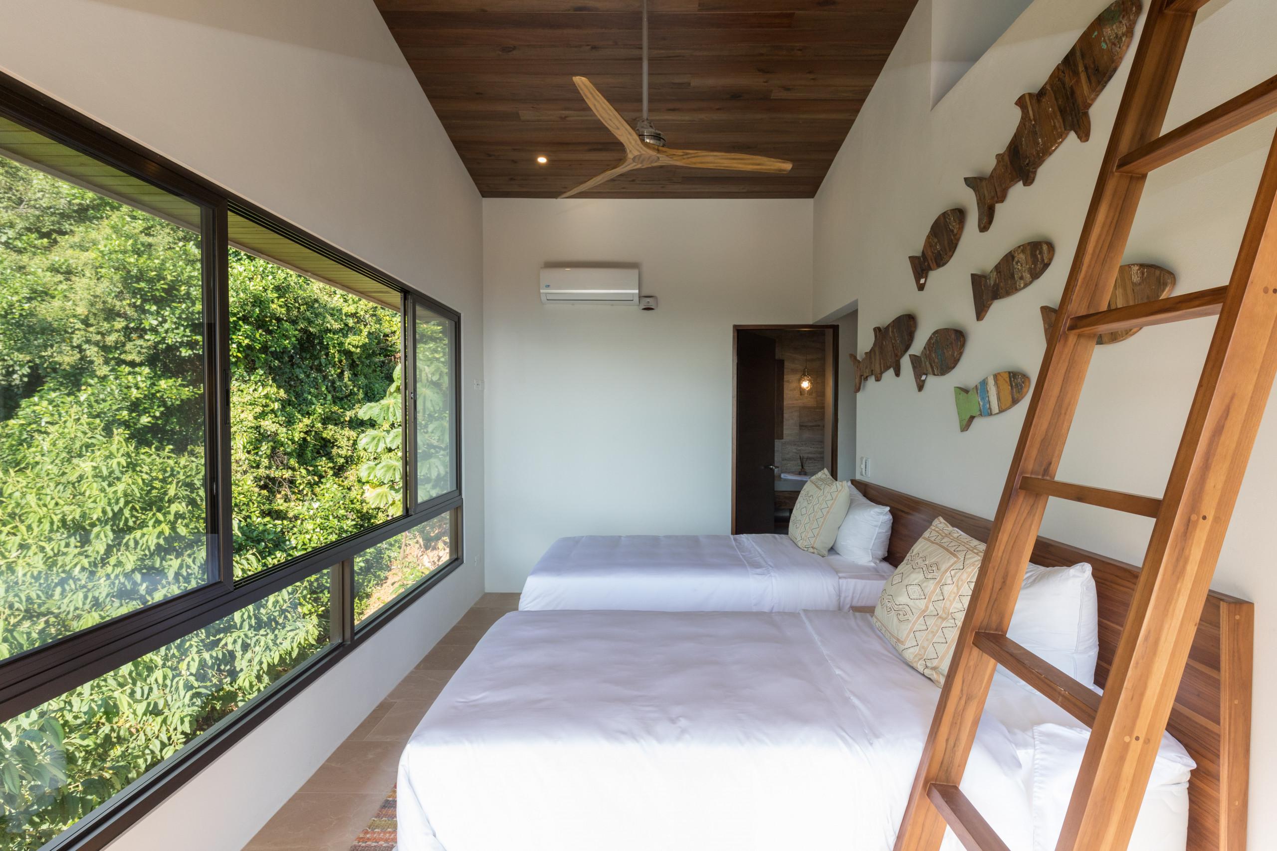 This modern bedroom features two single beds, a warm wood ceiling, and a wall of windows that invite natural light and jungle views.
