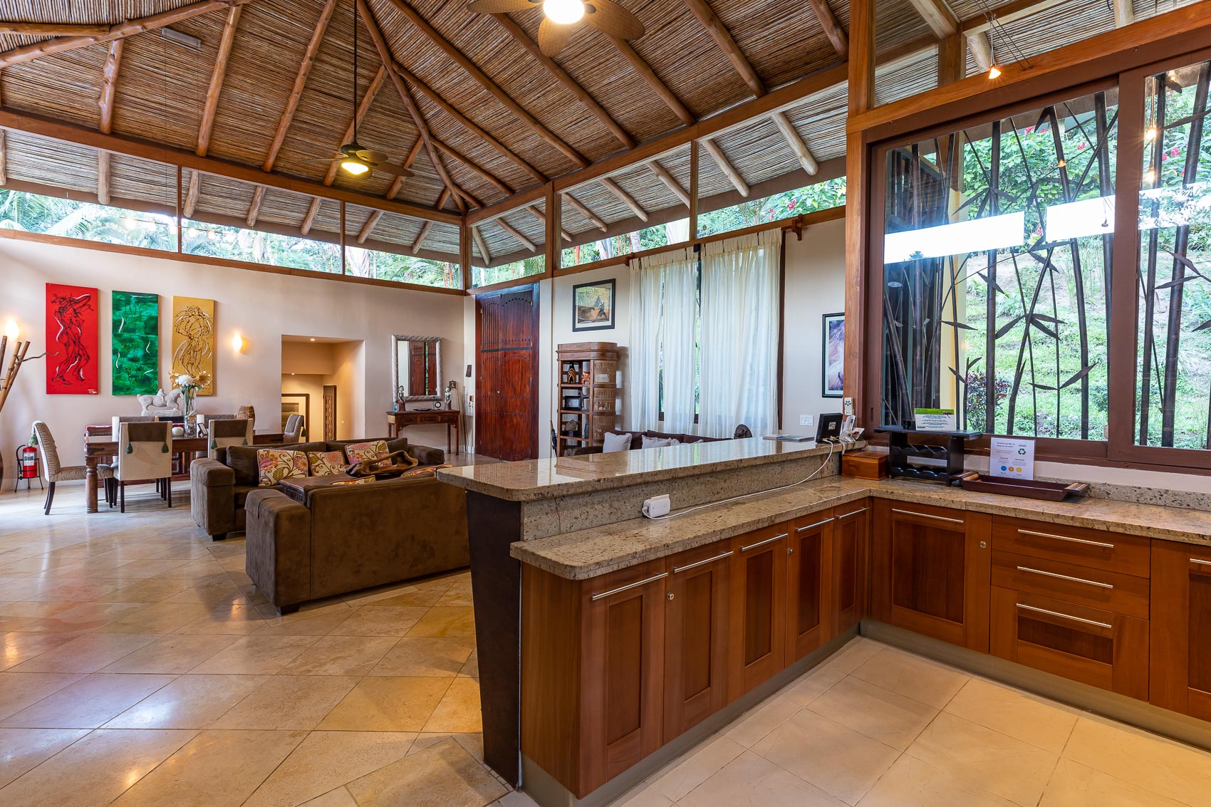 The expansive open-plan living area, connected to a dining space and kitchen counter, features a high vaulted bamboo ceiling, abundant natural light, and views of lush tropical greenery.