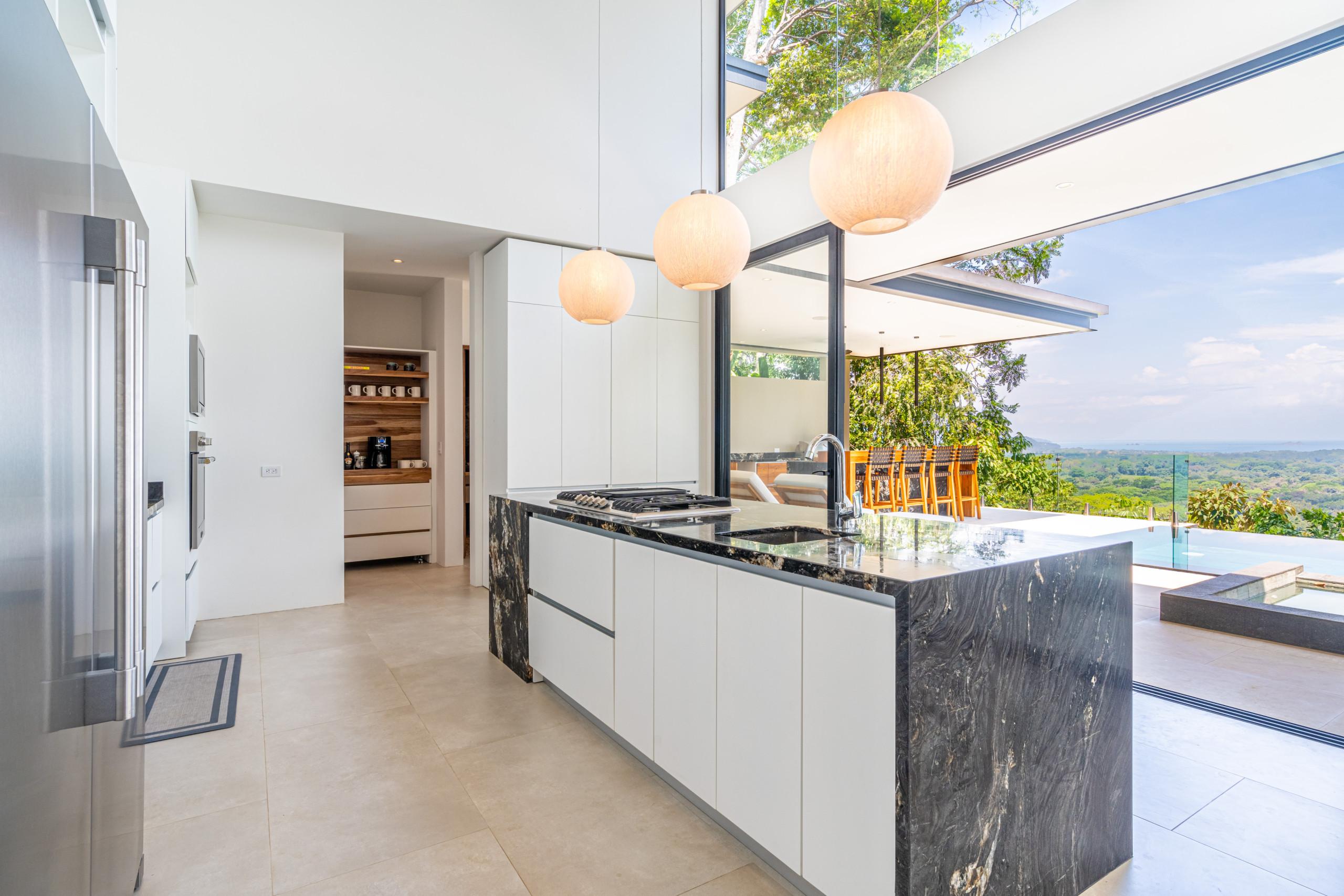 This modern kitchen features a large island with an integrated stovetop and sink, built-in appliances, and seamlessly opens to an outdoor terrace offering panoramic ocean and jungle views and access to an infinity pool.