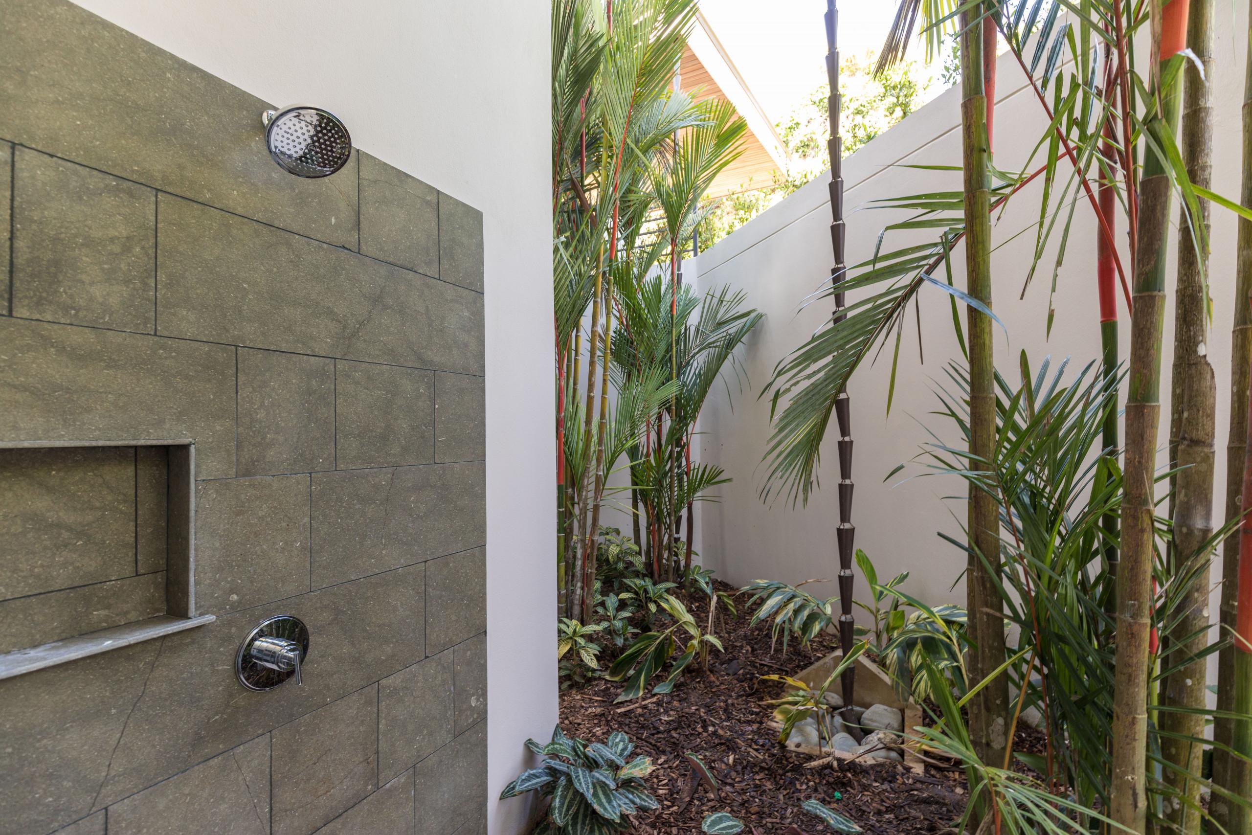 A stone-tiled outdoor shower with a built-in niche is situated alongside a private tropical garden featuring tall bamboos and lush undergrowth, bathed in natural light.