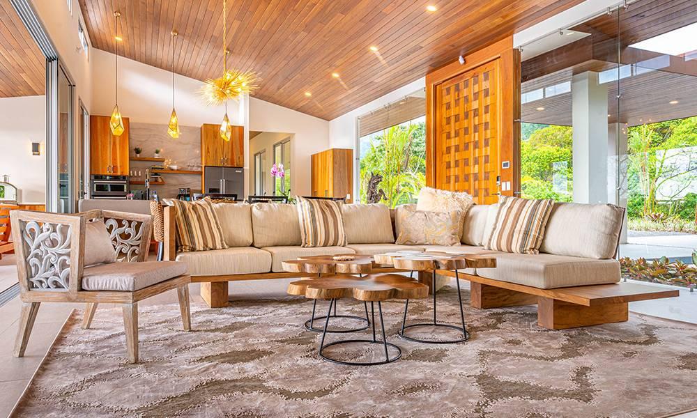 An open-concept living room with a sloped wood-paneled ceiling and a large sectional sofa connects to an adjacent kitchen, with full-height glass walls offering views of the surrounding jungle.