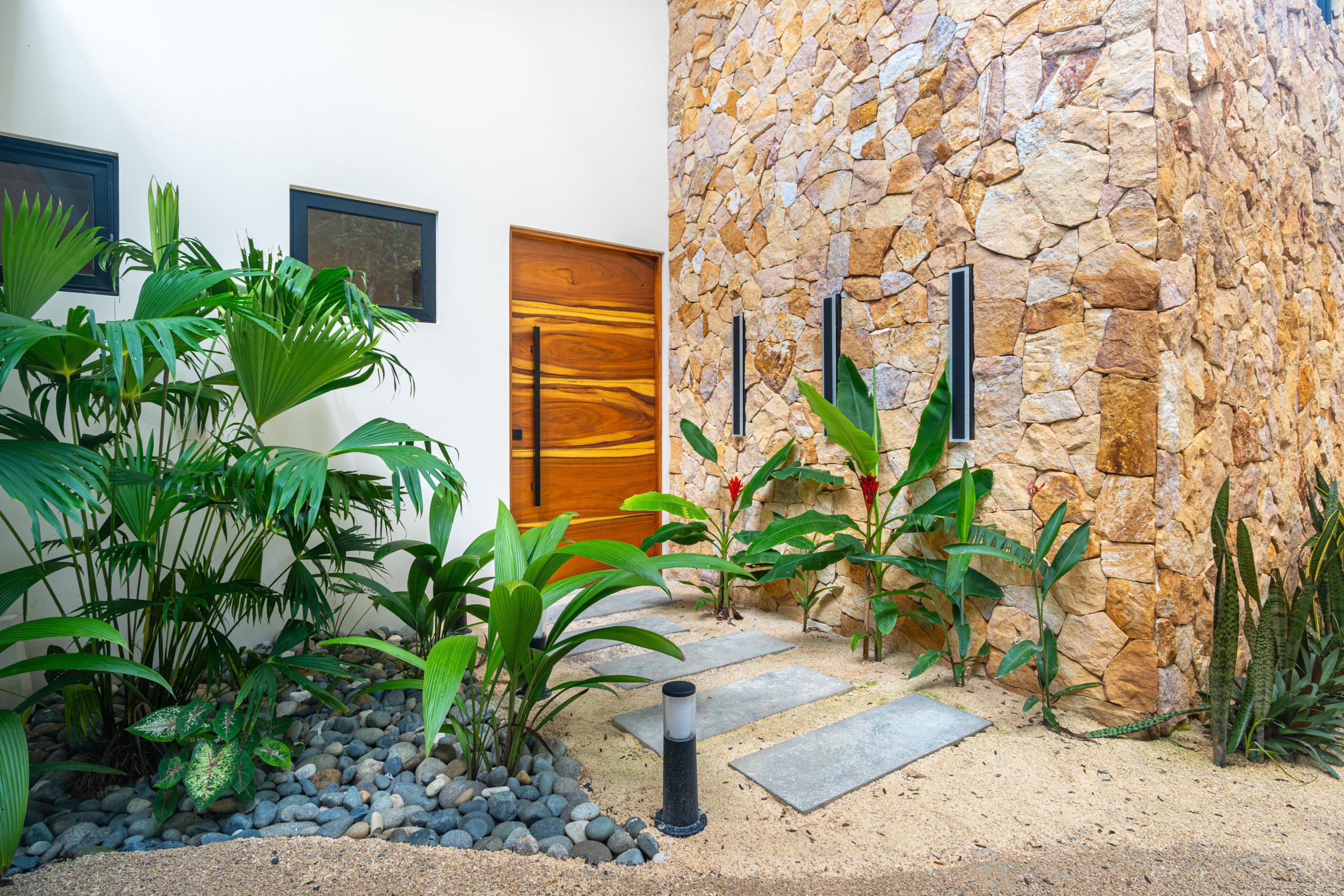 The villa entrance features a prominent wooden door, framed by a natural stone wall, a clean white exterior with windows, and lush tropical plants along a path of concrete stepping stones.