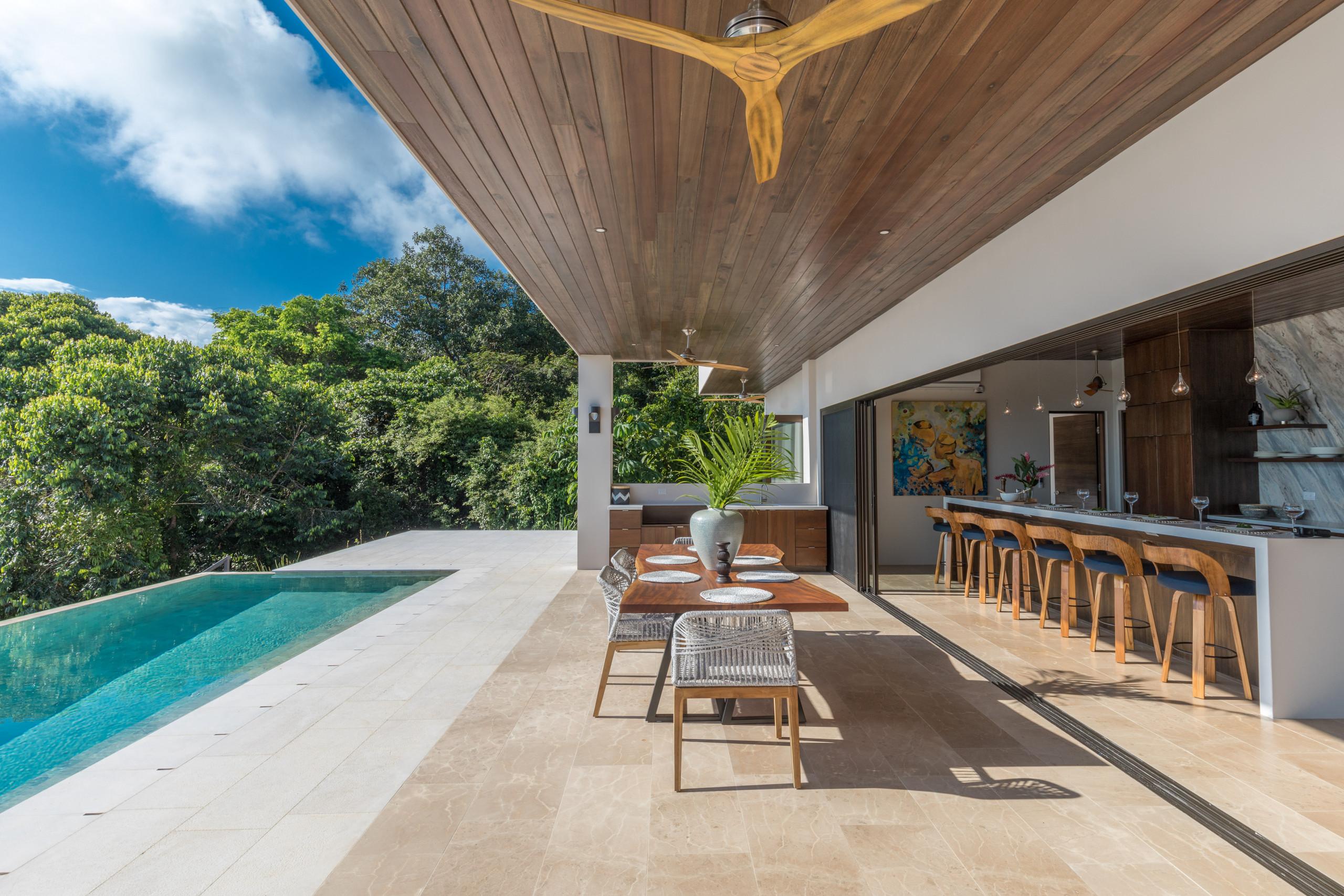 An open-air covered terrace with a wood-plank ceiling offers an infinity pool and outdoor dining, connecting to an indoor kitchen and jungle views.