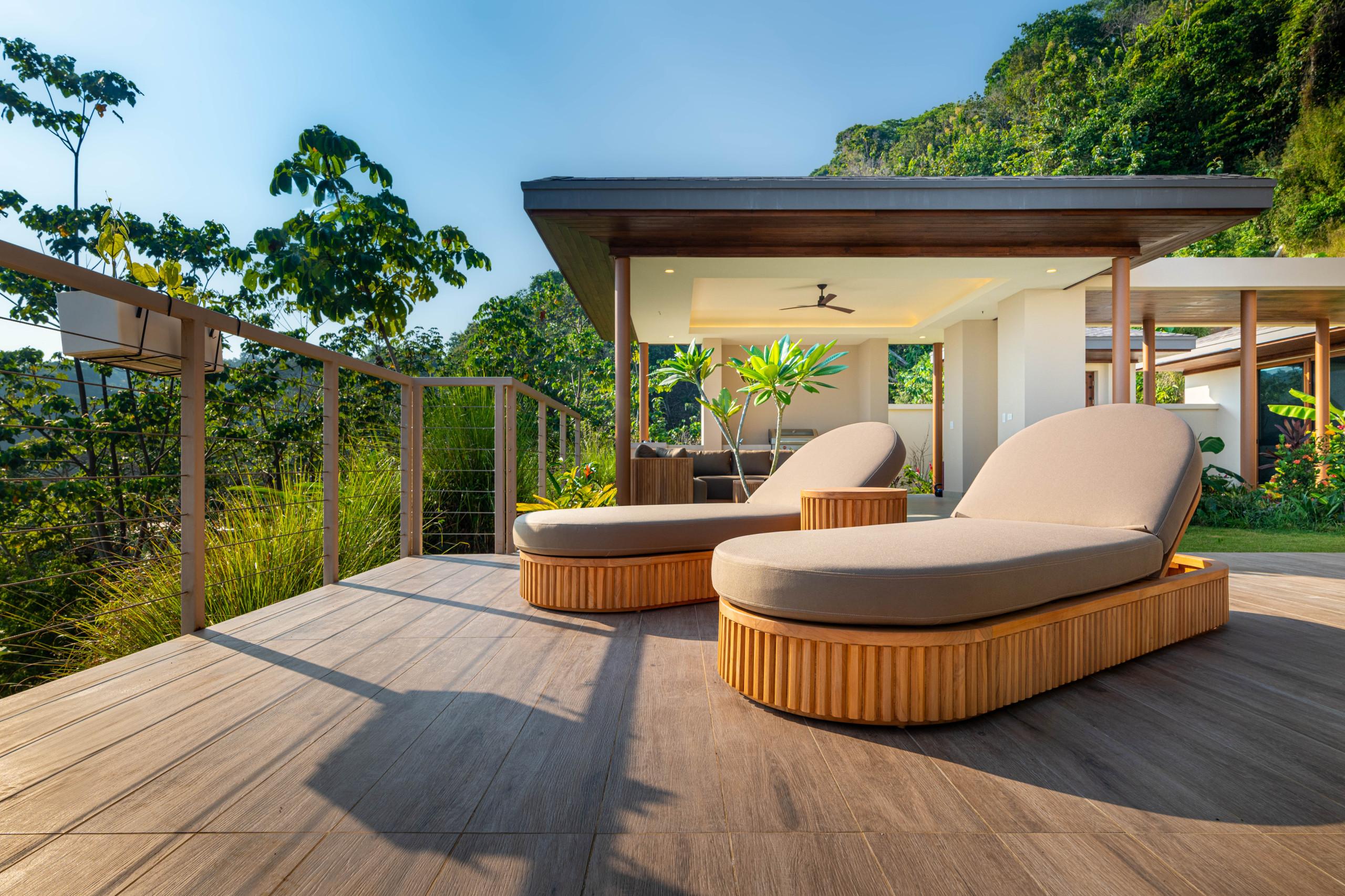 A sunlit wooden terrace features lounge chairs and a covered outdoor living space, opening to lush rainforest views and a clear blue sky.
