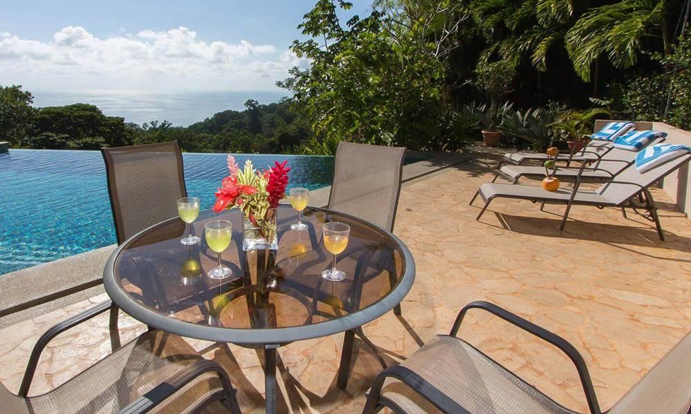 The outdoor area features an infinity pool with clear ocean and jungle views, alongside a flagstone deck with lounge chairs and a dining table.