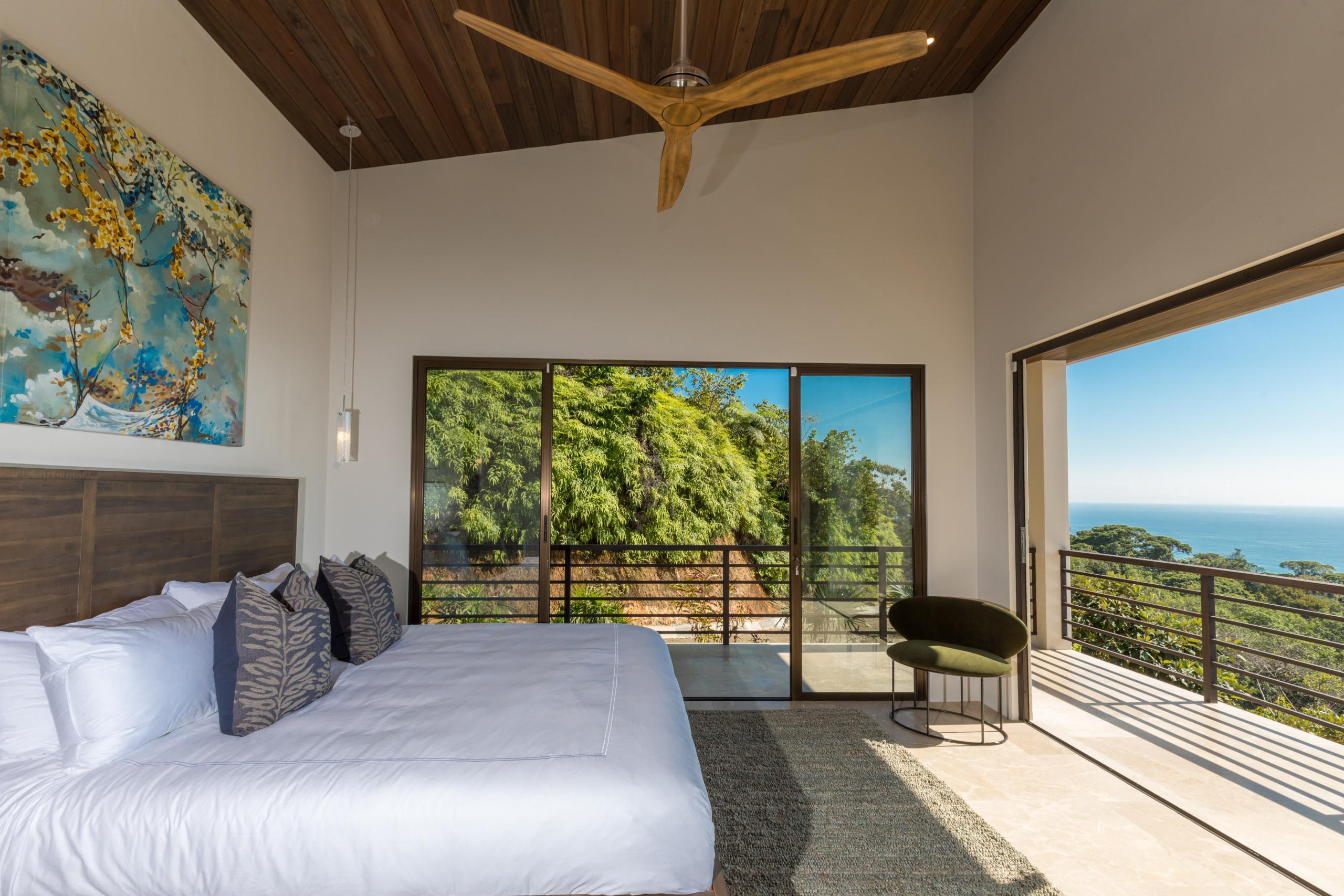This bedroom features a high wooden ceiling, a king-size bed, and extensive glass doors opening to a terrace with views of the jungle and distant ocean.