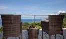A private terrace features two wicker chairs and a small table overlooking the expansive blue ocean and distant green coastline, bathed in natural sunlight.