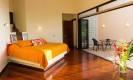 A spacious bedroom with rich wooden floors features glass walls opening to a balcony with views of the surrounding jungle, filled with natural light.